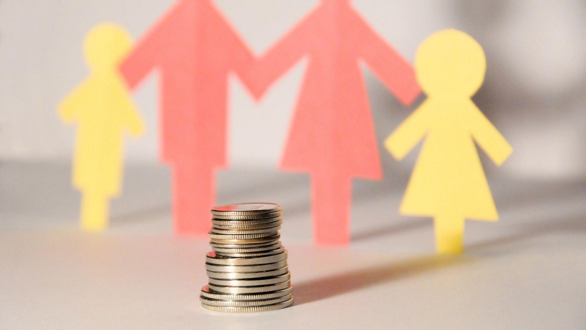 A stack of coins sits in the foreground of the photograph. Behind, slightly blurred in the background, are paper cutouts of people. Two adults are in red with two yellow children either side. 