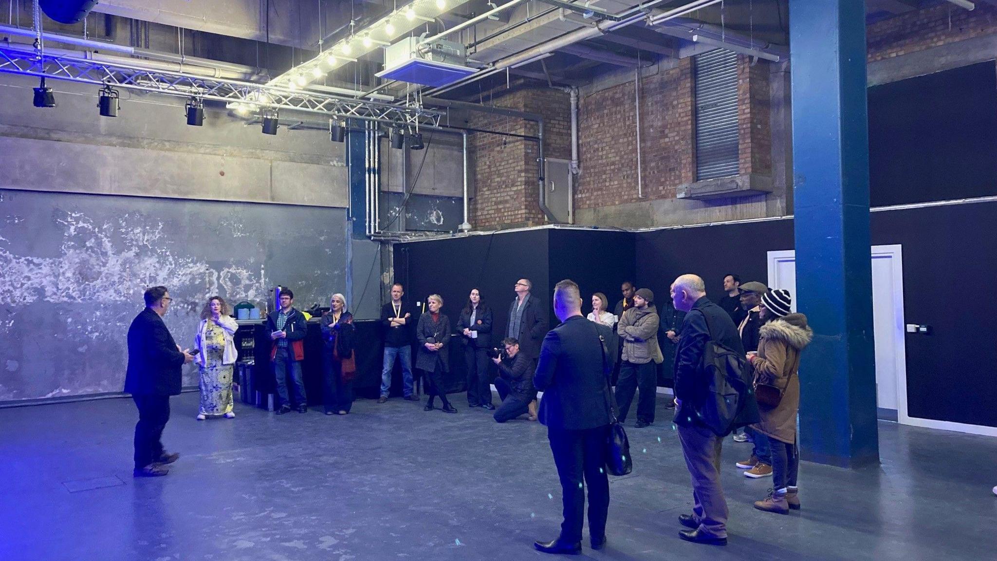 A group of people standing in a circle inside a large, industrial brick-walled room with metal lighting racks. 