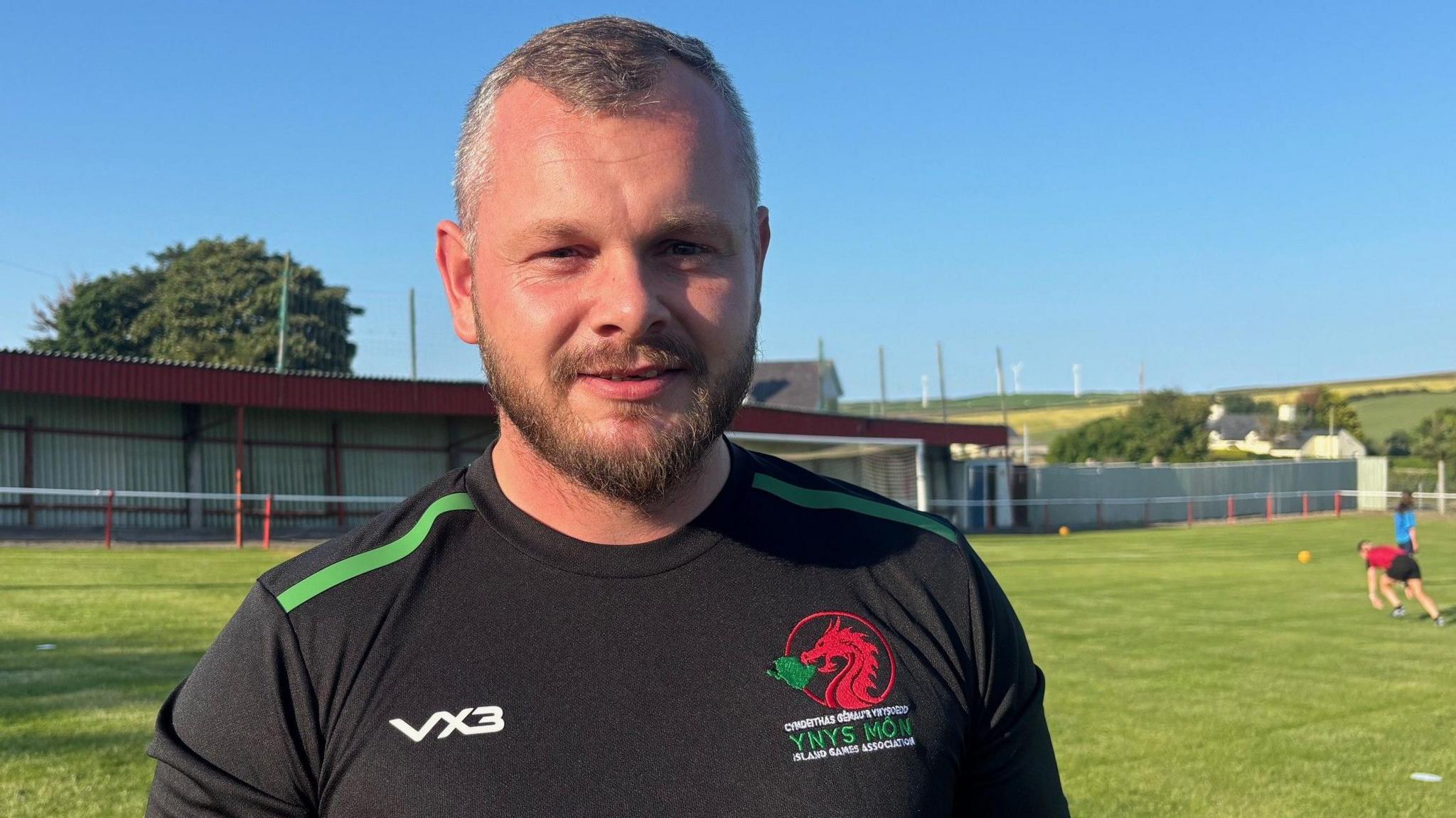Llun o Liam yng ngolau'r haul yn sefyll ar gae pêl-droed Cemaes. Mae rhai o chwaraewyr y tim yn ymarfer y tu ol iddo, ac mae o'n gwisgo crys pêl-droed du gyda gwyrdd a choch arno. Mae ganddo wallt byr ac mae'n gwenu ar y camera.