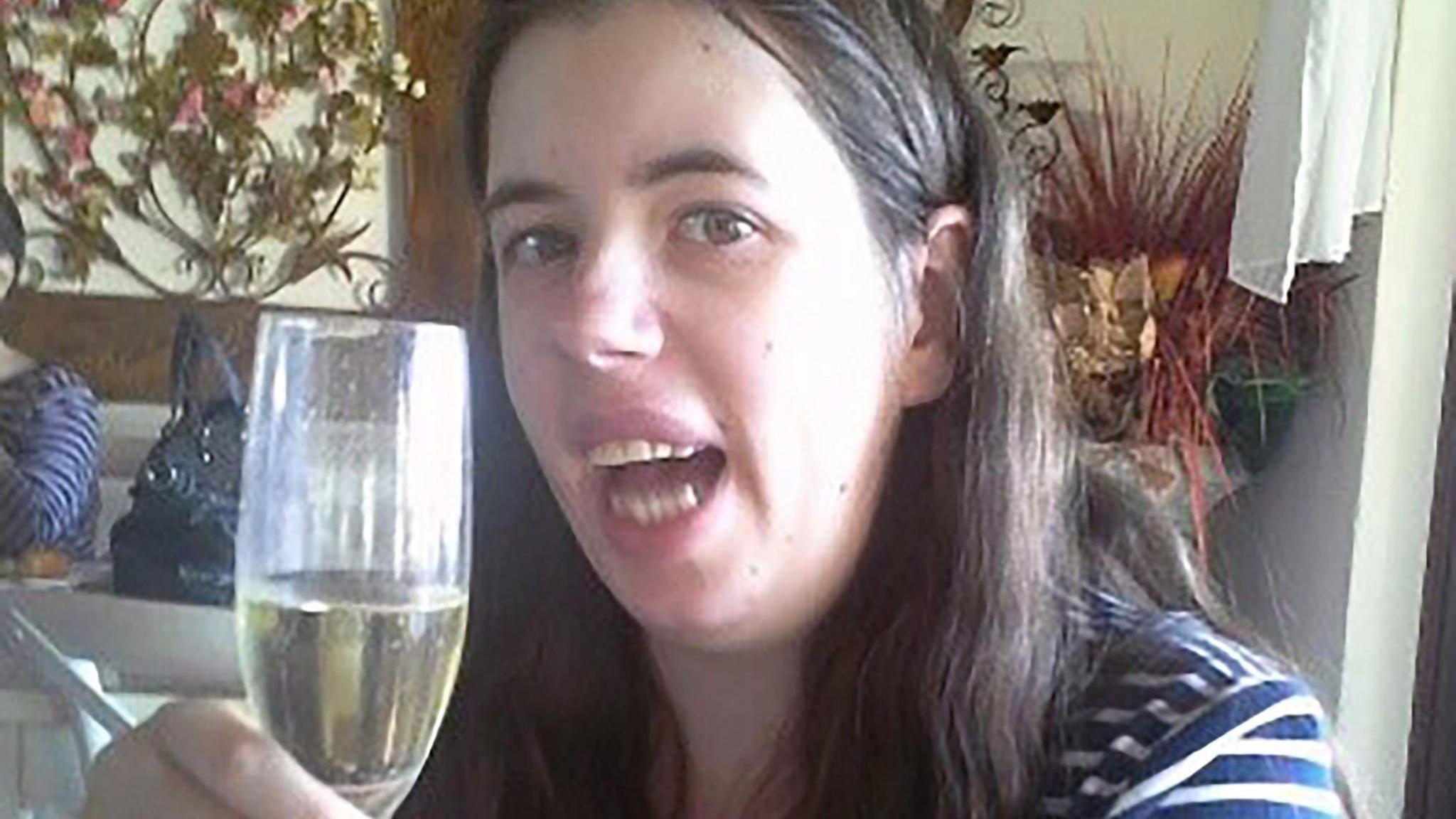 Chloe Every pictured holding a champagne flute with a sparkling liquid inside. She appears to be in a restaurant or similar environment, with tables and flowers. She is wearing a navy and white striped top and is looking toward the camera as if making a toast.