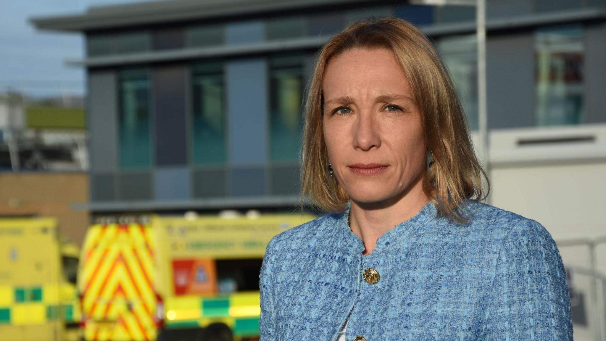 A woman wearing a light blue jacket, standing in front of two ambulances outside a hospital