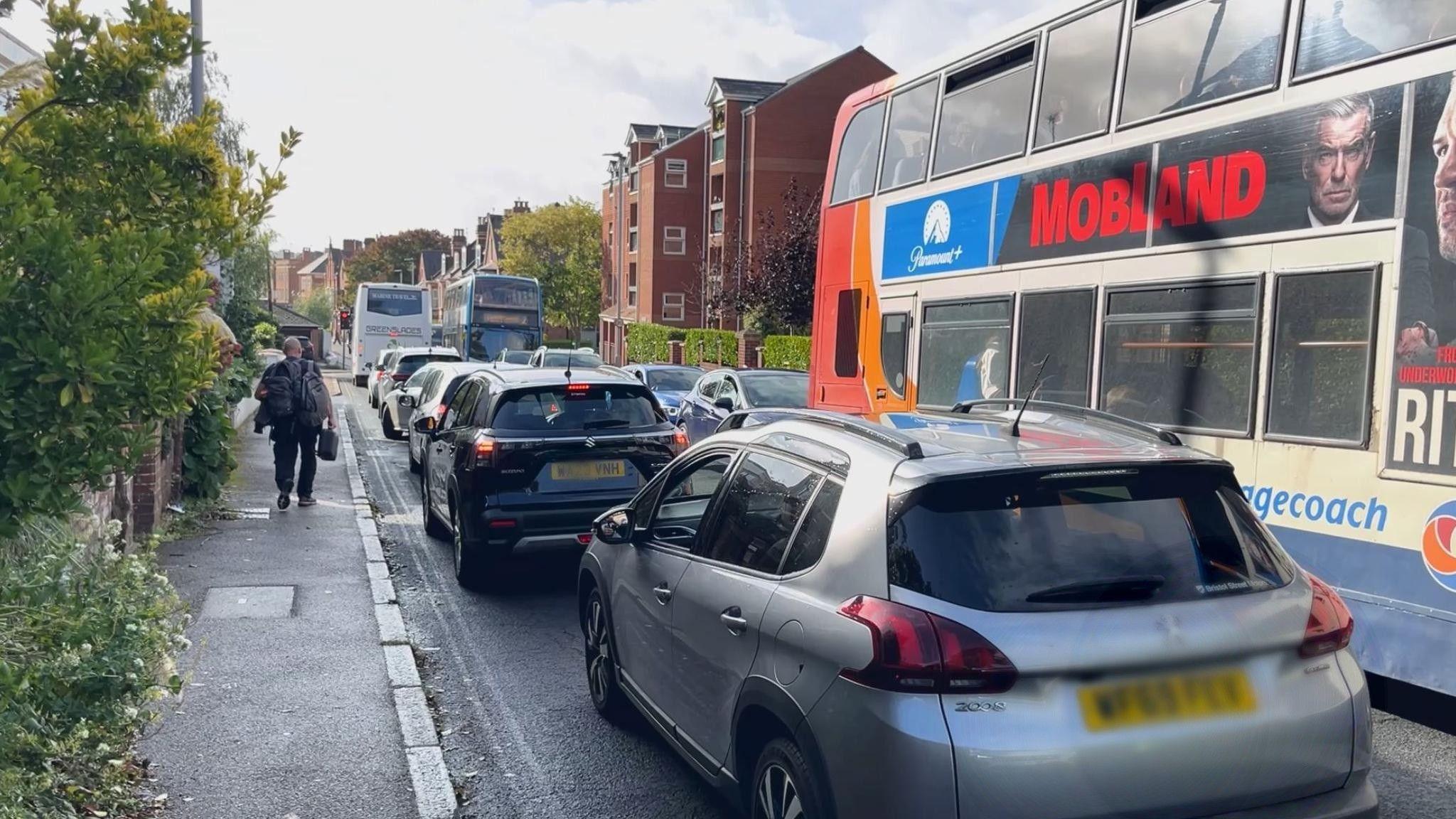 Cars queuing going towards Exeter city centre along Magdalen Road and the side of a bus with several cars queuing behind going in the other direction. A man is walking along the pavement on the left.