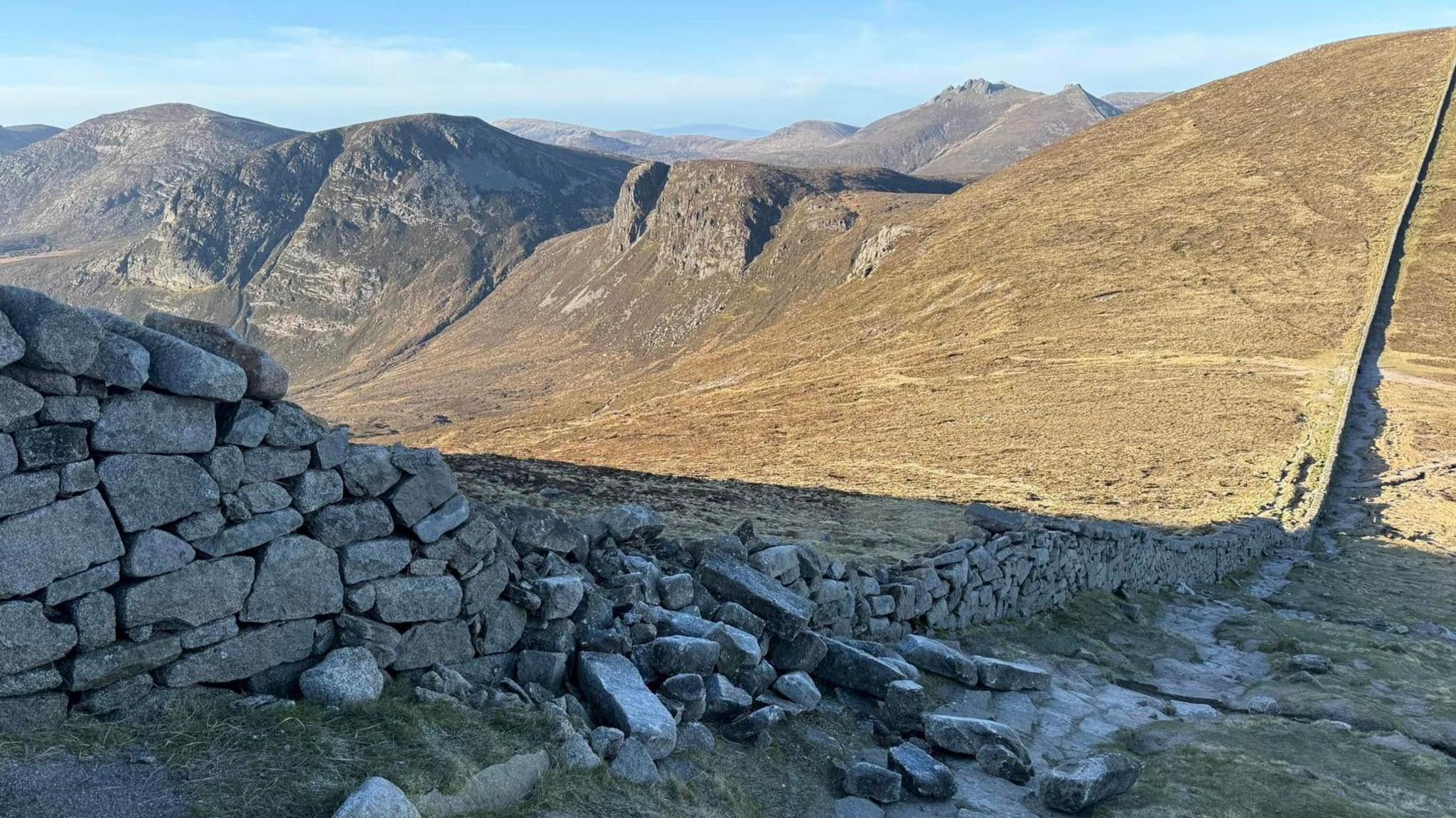 A granite stone wall slopes down the side of mountain. A section of the wall has collapsed and a large number of stones are lying in rubble.