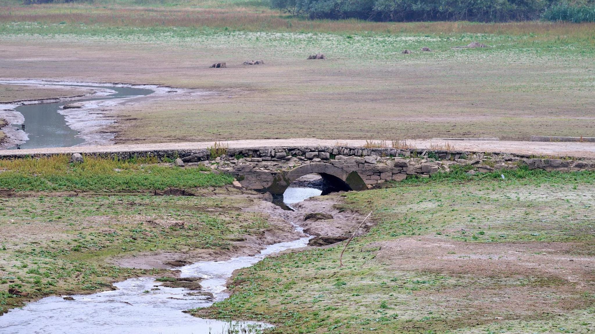 An old arched bridge and a road crossing low lying grassland. There is a small stream running under the bridge.