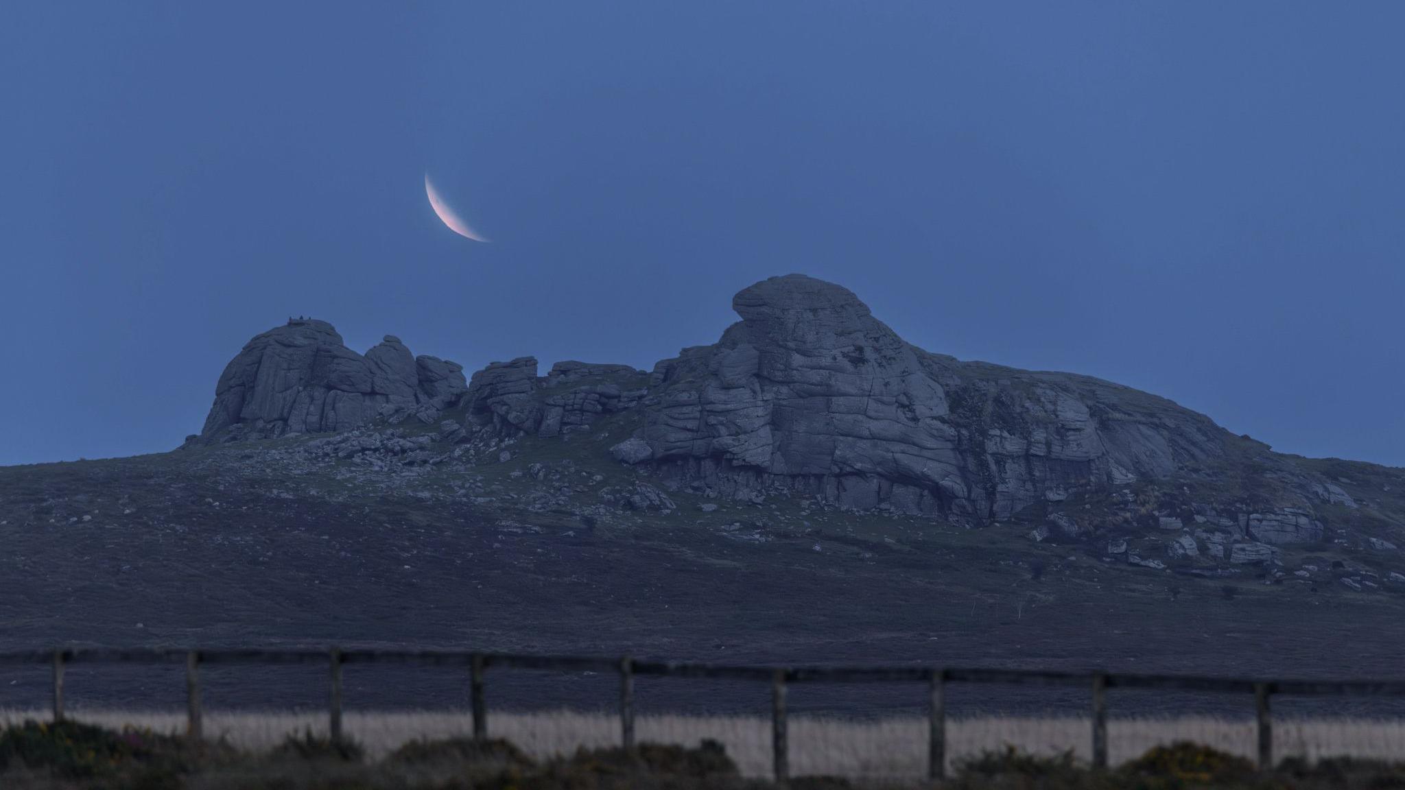 The moon rising over Haytor, the tor, a large rocky outcrop juts out from the landscape. There is a low fence in the foreground. The sky is dark blue. 