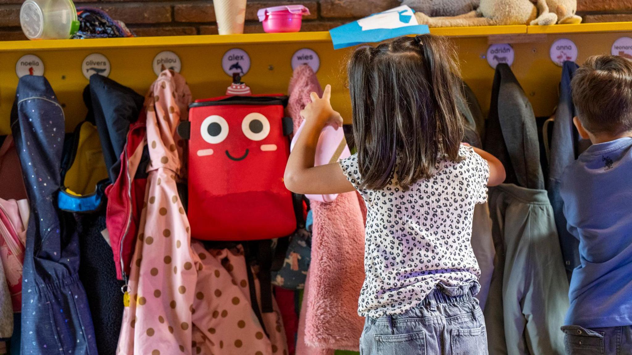 A young child hangs up her coat on a row of pegs labelled with children's names.