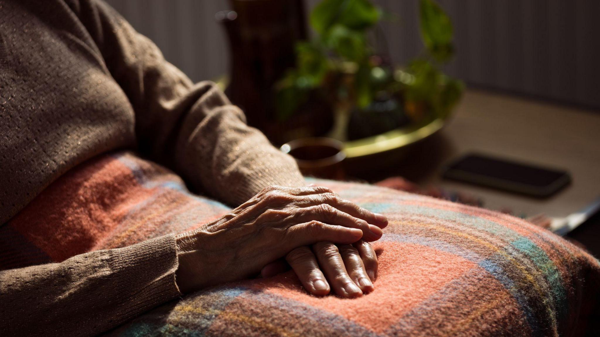An older woman sitting in a chair with an orange and blue blanket on her lap. Her hands are on top of one another on her lap. She is wearing a brown jumper.