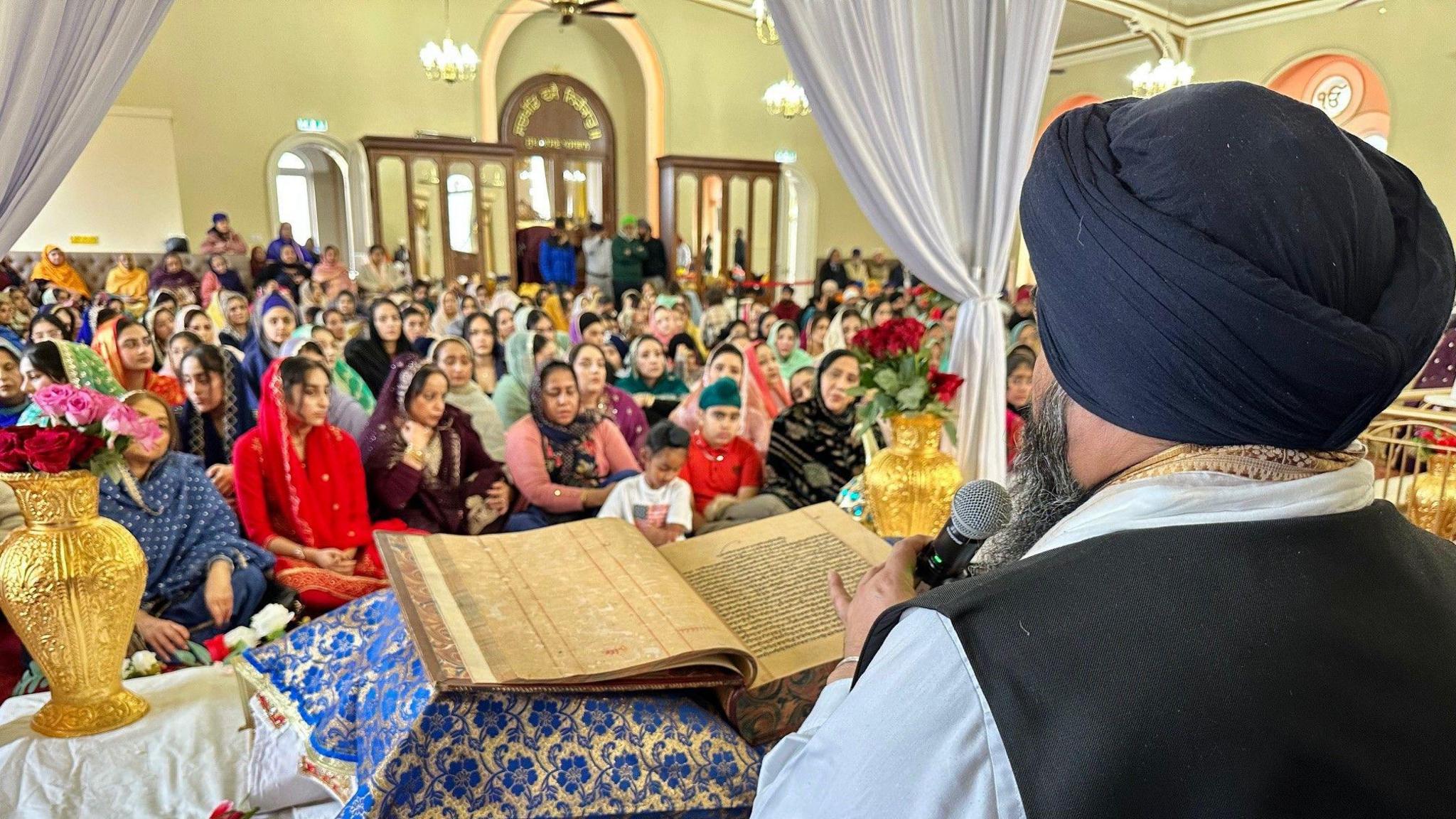 The picture is taken behind the priest. the book is in front of him and the crowd is in the background.