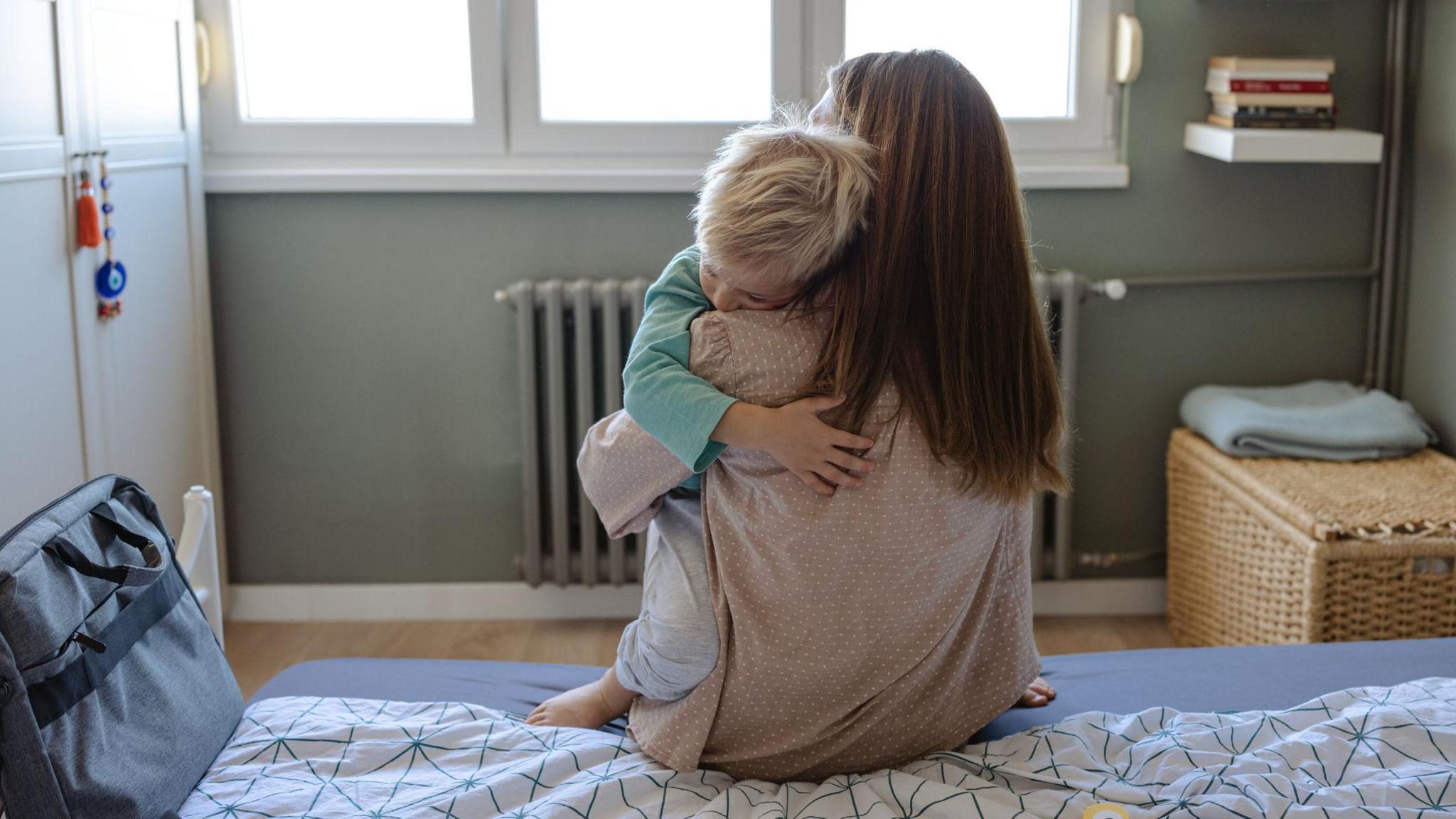 A mother embraces her child while seated on a bed in a bedroom.