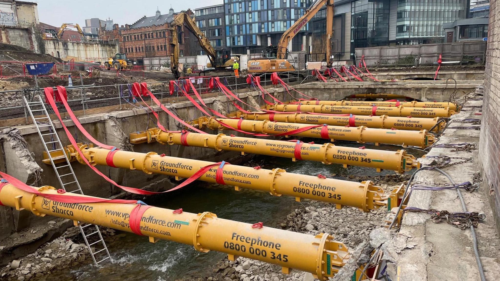 Several large yellow tubes stretch out high above a riverbed from one bank to another. The river below is shallow and lined with rocks