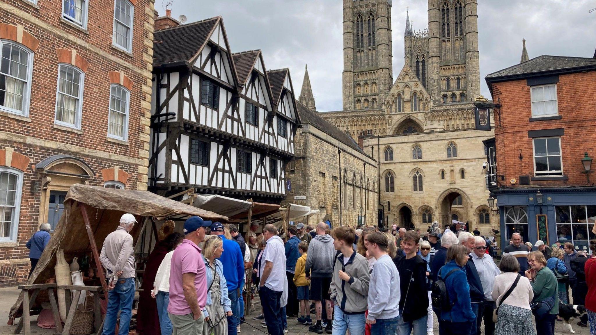 Crowds of people wander around a market in a square surrounded by historical buildings, including a Tudor-style house and a large medieval cathedral with three towers.
