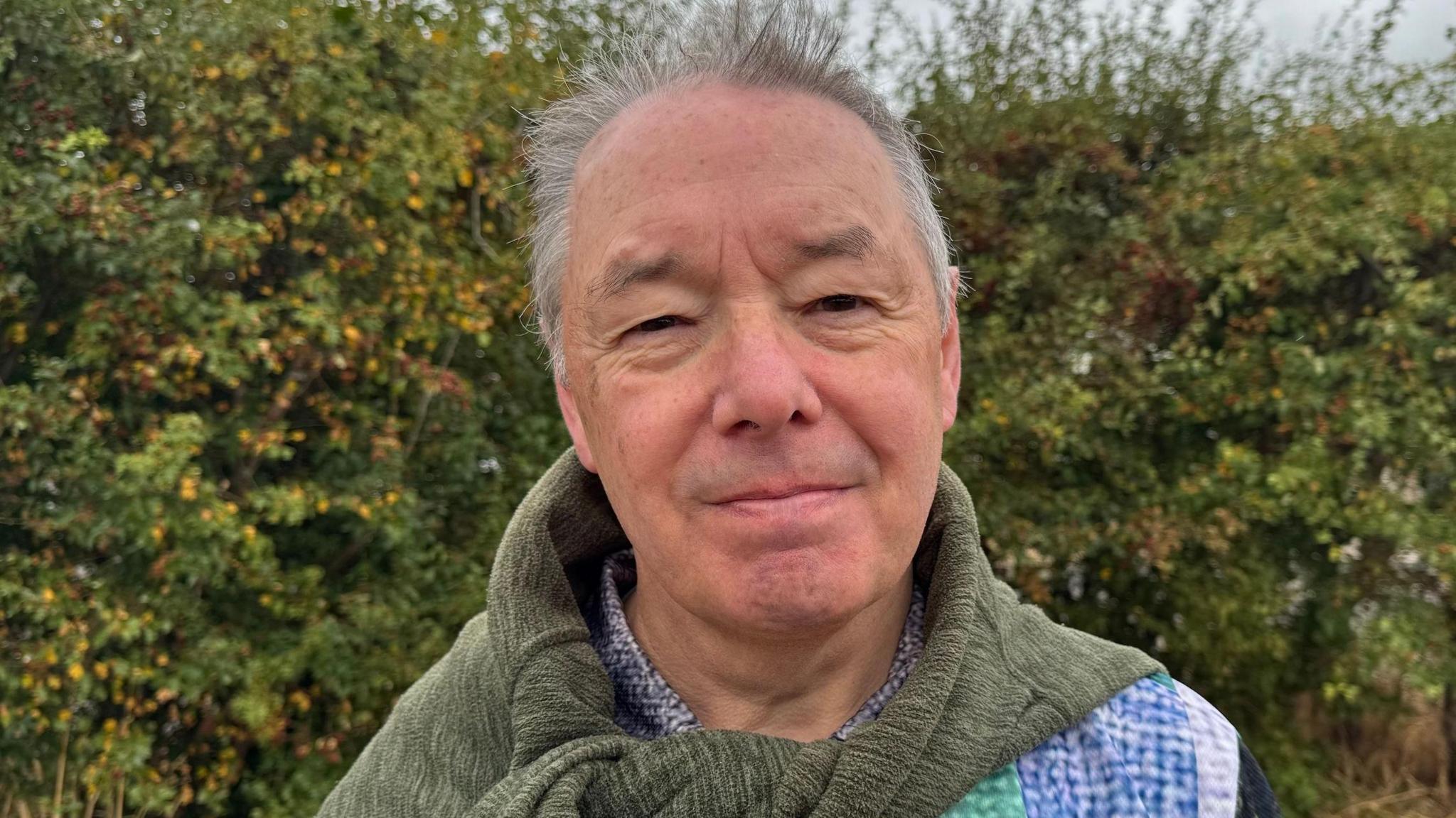 Mark Higgitt is wearing a blue-and-green checked shirt and he has a green jumper tied around his neck. He has short, grey hair, and he's looking at the camera.
