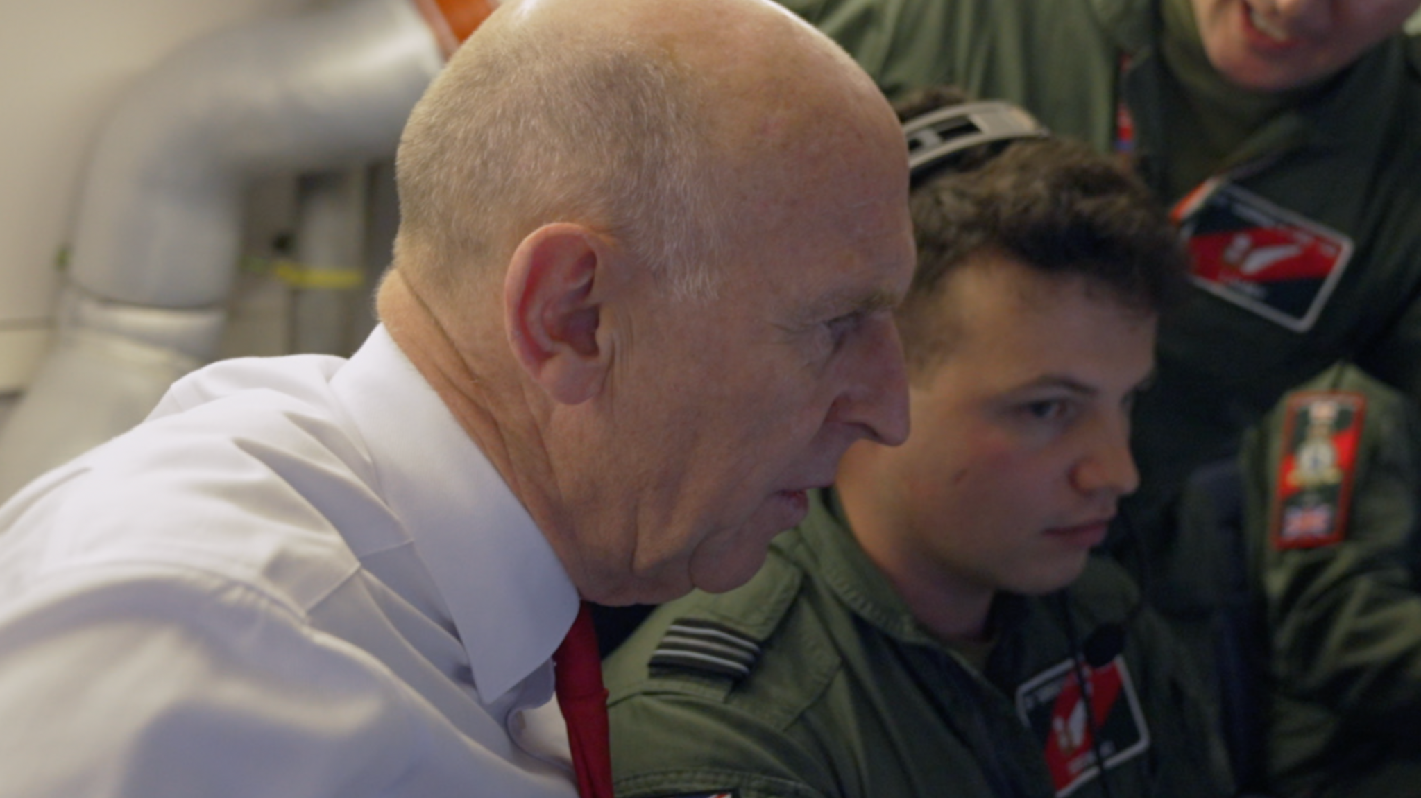 Defence Secretary John Healey in side profile, wearing a white shirt and red tie, alongside RAF crew in uniform