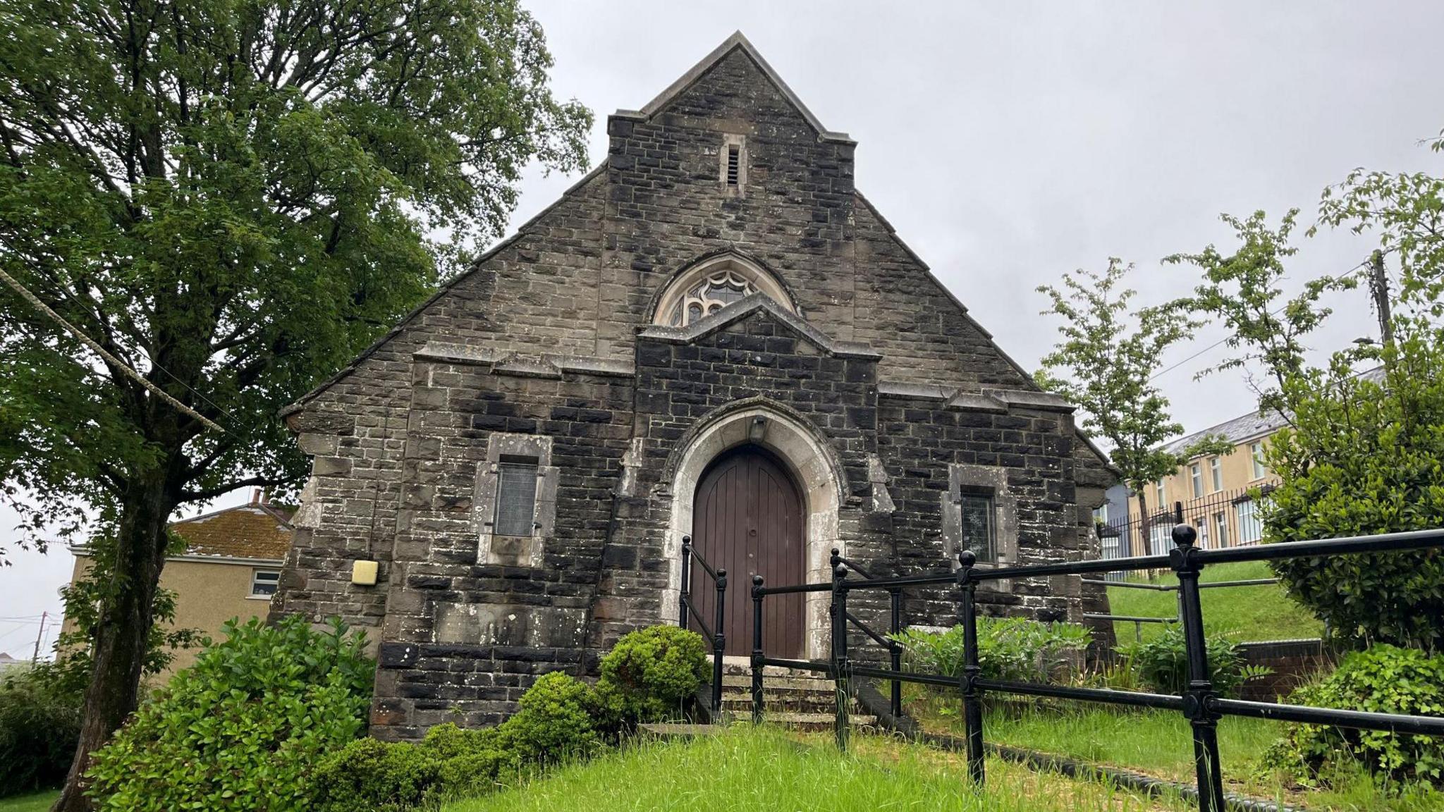 Outside of Markham Congregational Chapel. A small grey stone chapel, on the side of a hill. A large arched wooden door is at the middle of the chapel, with two narrow windows either side. It sits among some grass and a path with railings. Trees can be seen either side of the chapel, with a row of houses up the hill behind it.