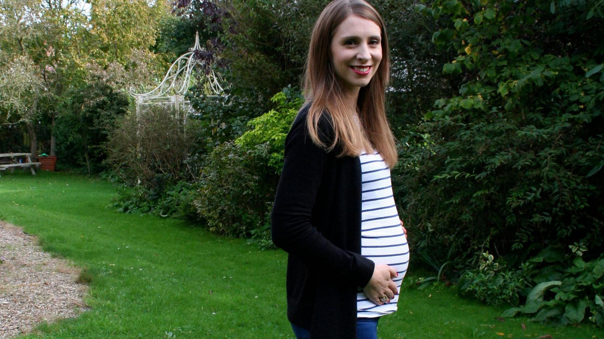 Ali Harris is standing outdoors in a peaceful garden setting. She’s wearing a black cardigan over a white and blue striped top, paired with blue jeans. Her long, straight hair frames her face, and she’s smiling at the camera. One hand rests gently on her stomach, she is pregnant. Behind her, the garden is full of greenery, there are trees, bushes, and a grassy lawn. A white, decorative metal structure and a gravel path are also in the background.