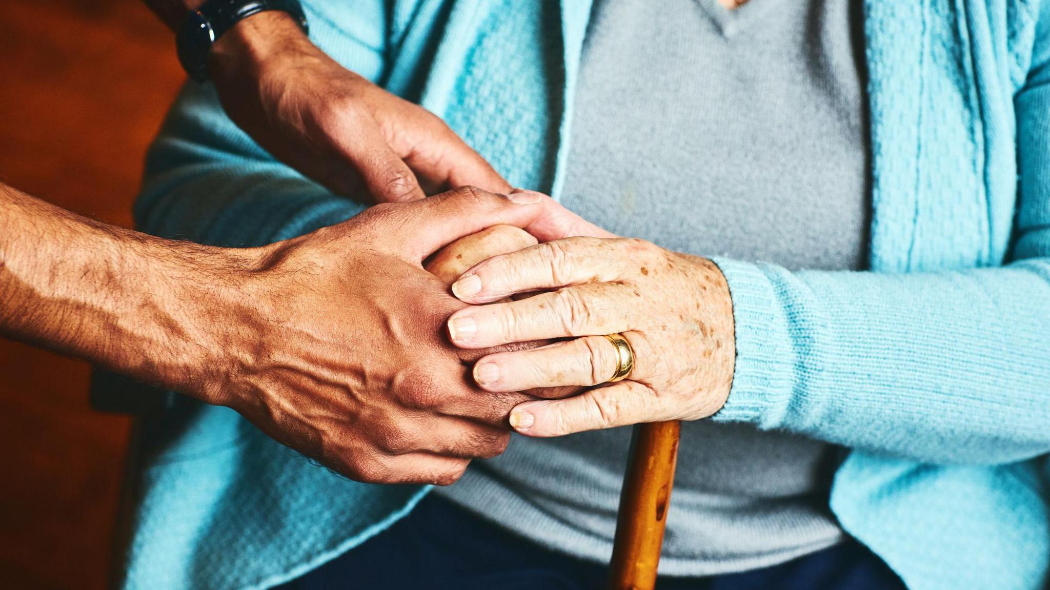 A pair of hands is supporting a person who is holding onto a brown walking stick. The person with the walking stick is wearing a blue cardigan.