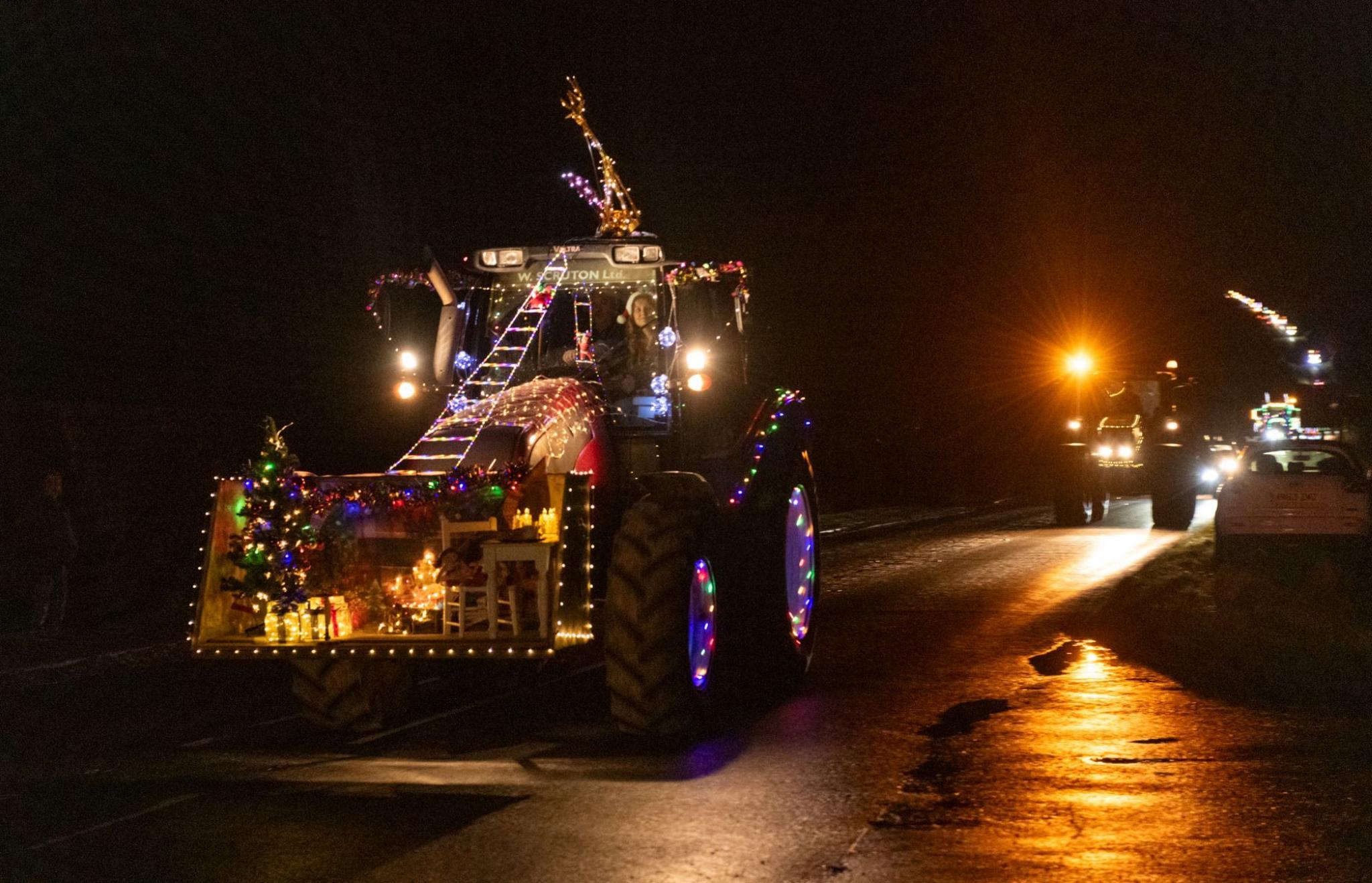 A tractor has a model living room scene attached the front. including a Christmas tree, presents, fireplace, tables and chairs.