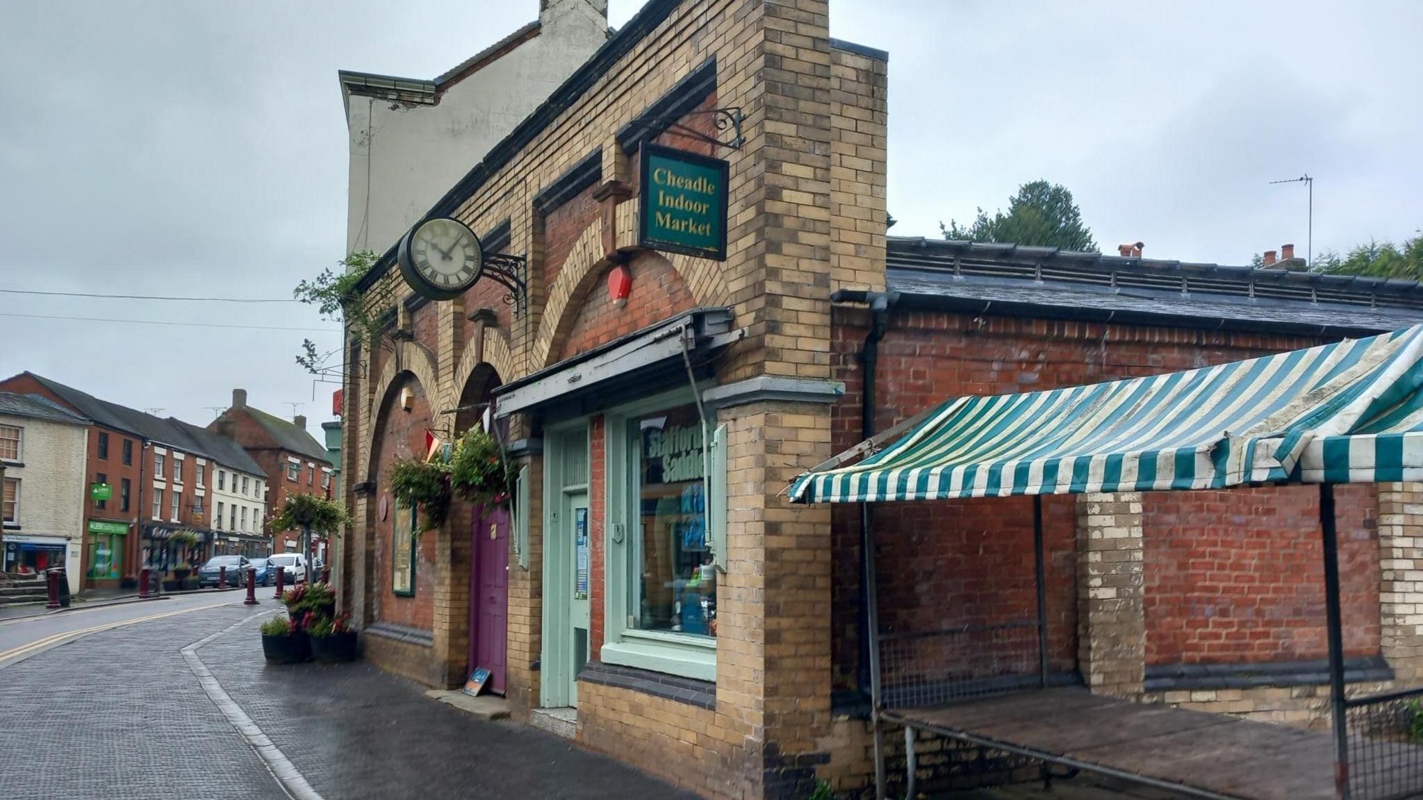 The front of a market hall building on a town's high street. A sign above it says Cheadle Indoor Market and there are flowers and a clock over the entrance. To the right, next to the building, is a table with a green and white market awning set up over it.