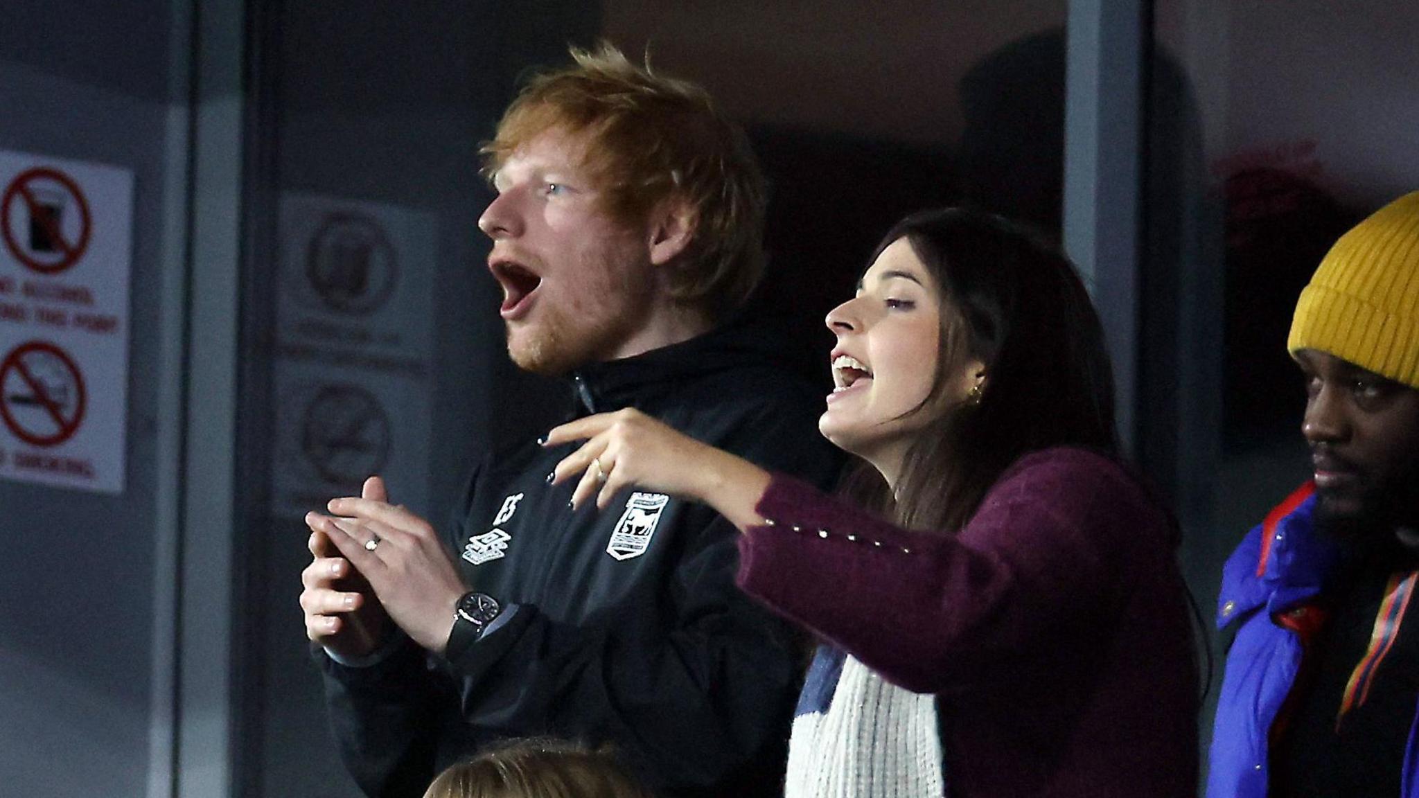 Singer Ed Sheeran and a young woman sing in support of Ipswich Town during the match with Manchester United at Portman Road. He is wearing a black top with the Ipswich crest on it, and she is wearing a plum-coloured coat with a large blue and white scarf