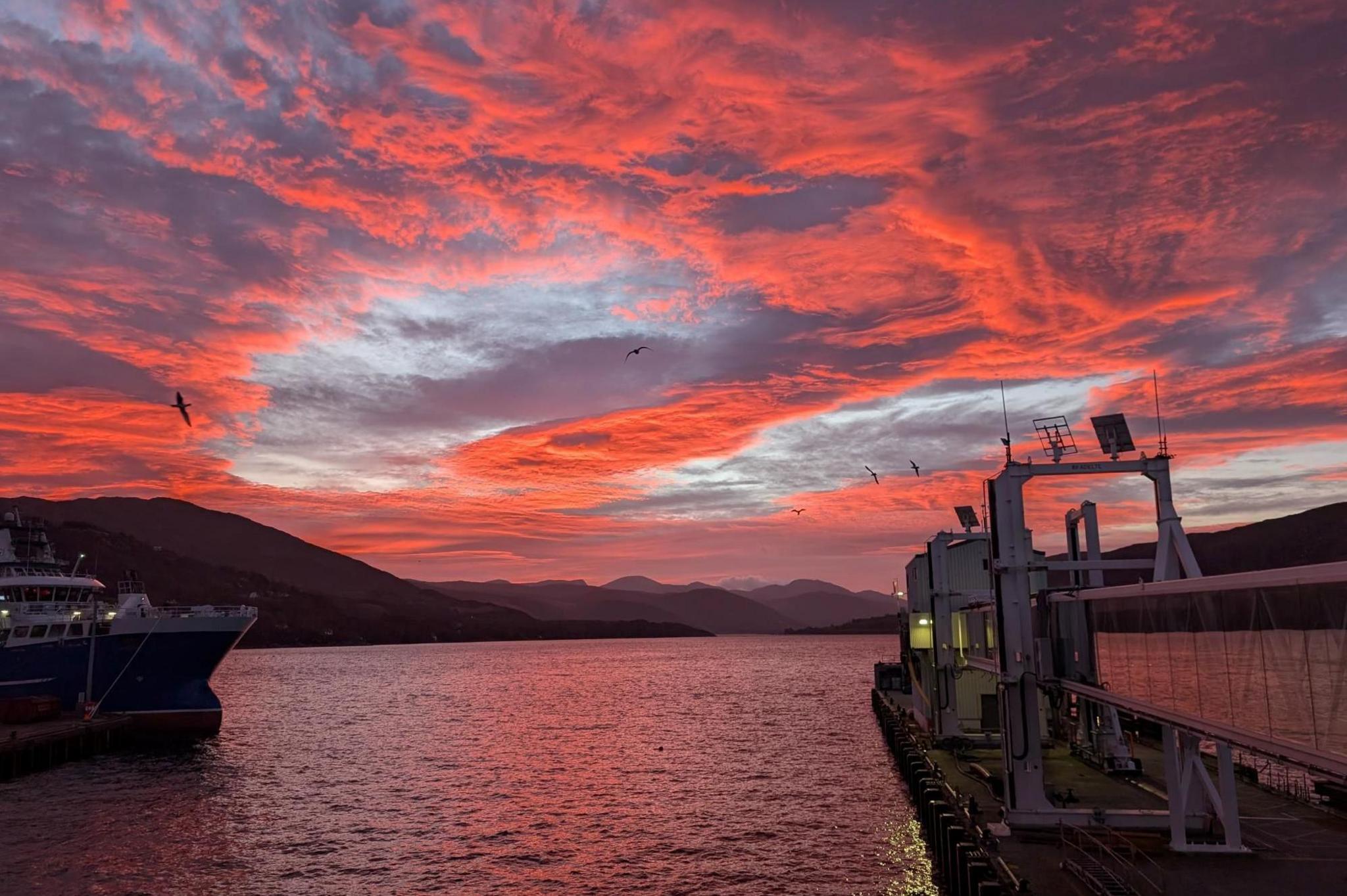 Clouds in the morning sky are glowing red. There is a fishing boat moored near the terminal, which stretches out into the sea.