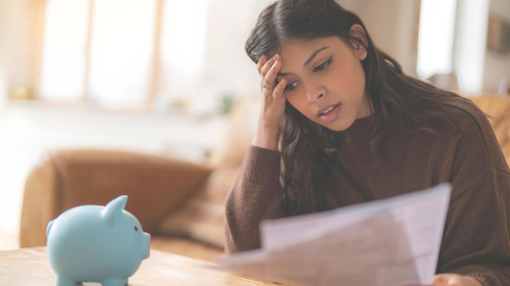 A young woman looks distressed as she holds a bill and looks at her piggy bank.