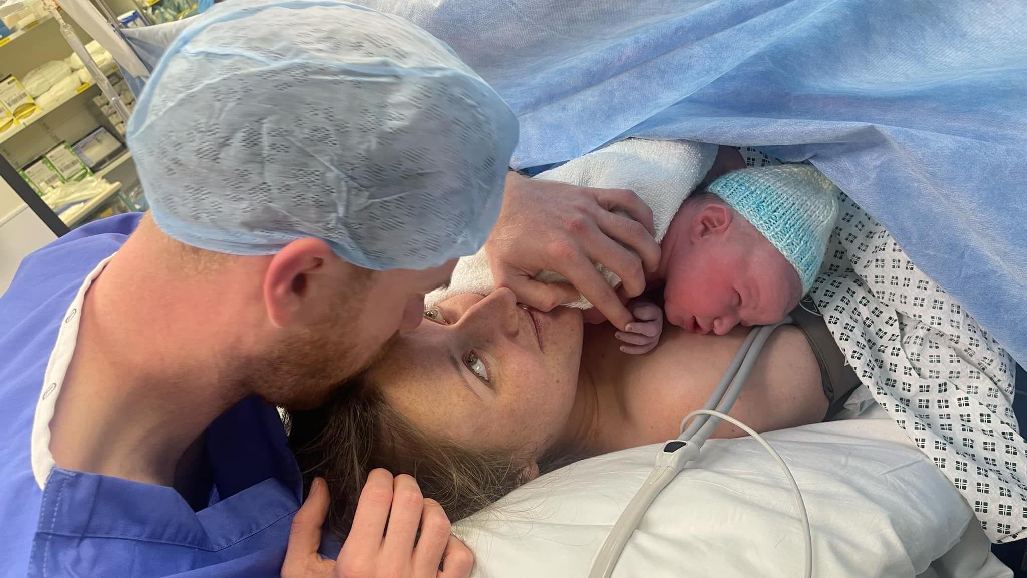 A woman in a hospital bed covered in a blue sheet with tubes attached holds her new born baby, who is wearing a knitted hat. Her partner kisses her head. He is wearing a blue hair net and blue hospital scrubs. 