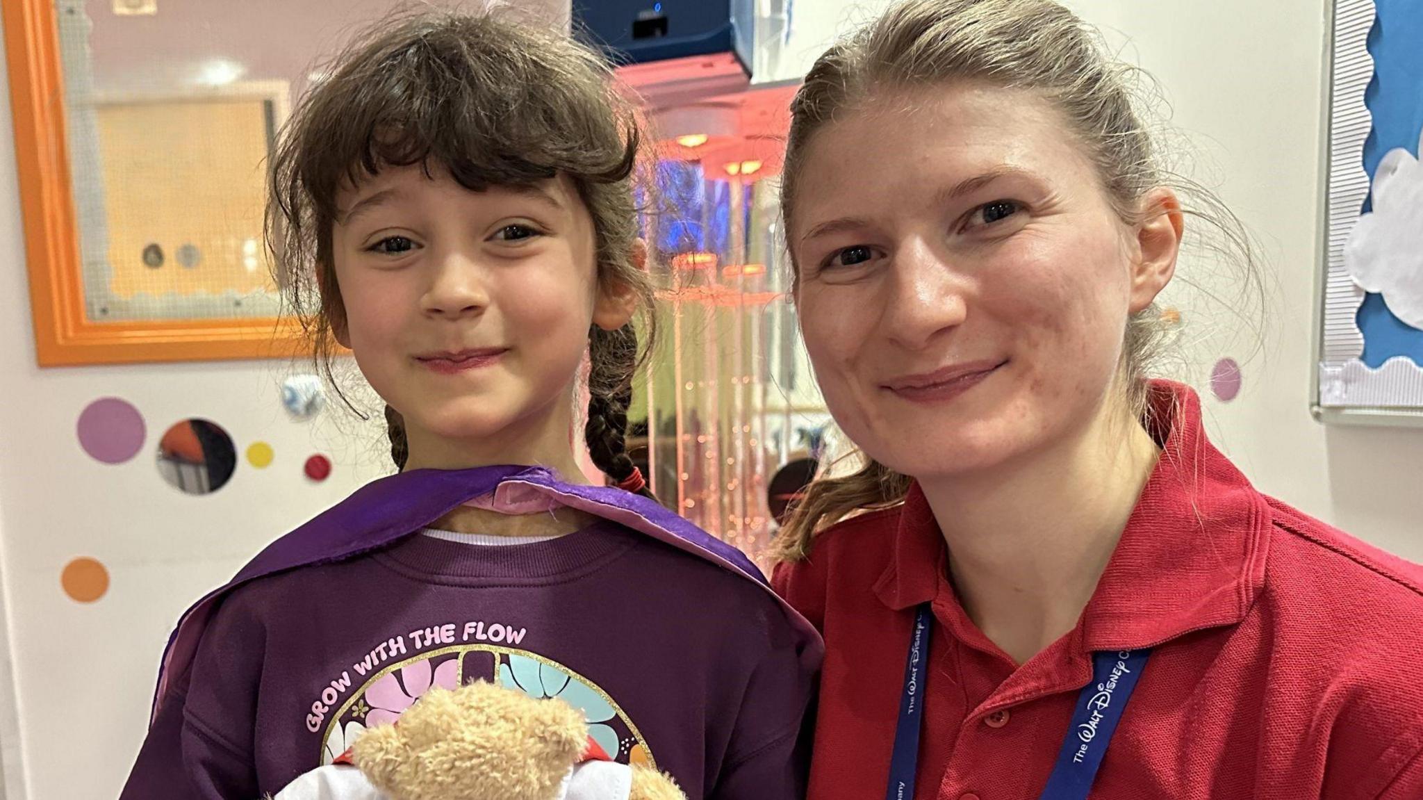 Sylvie with Play Specialist Naomi Taylor - they are smiling for the camera in the hospital. Sylvie is holding a small beige teddy bear. A play area could be seen behind them.