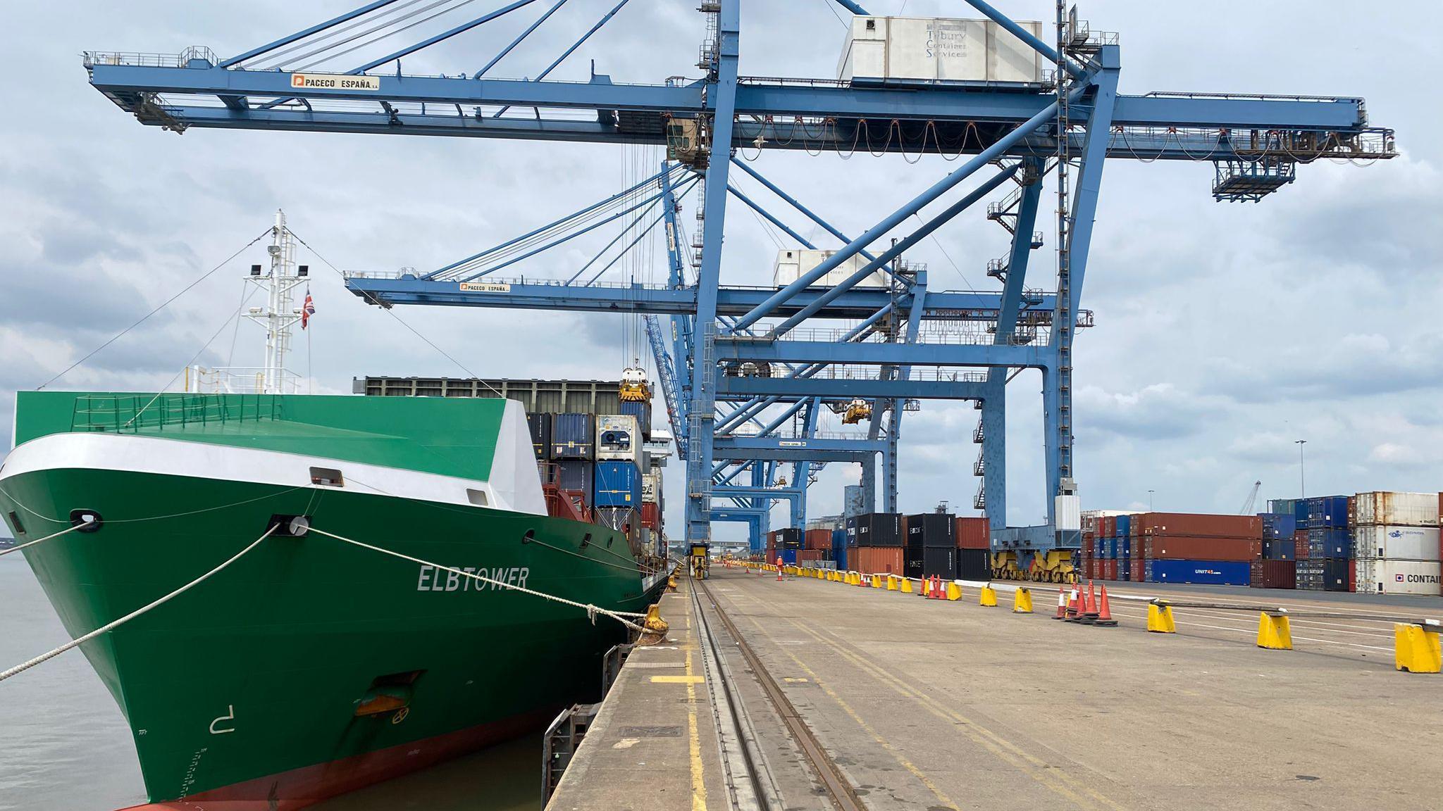 A green boat in the water with a large blue crane and multiple containers in the background.