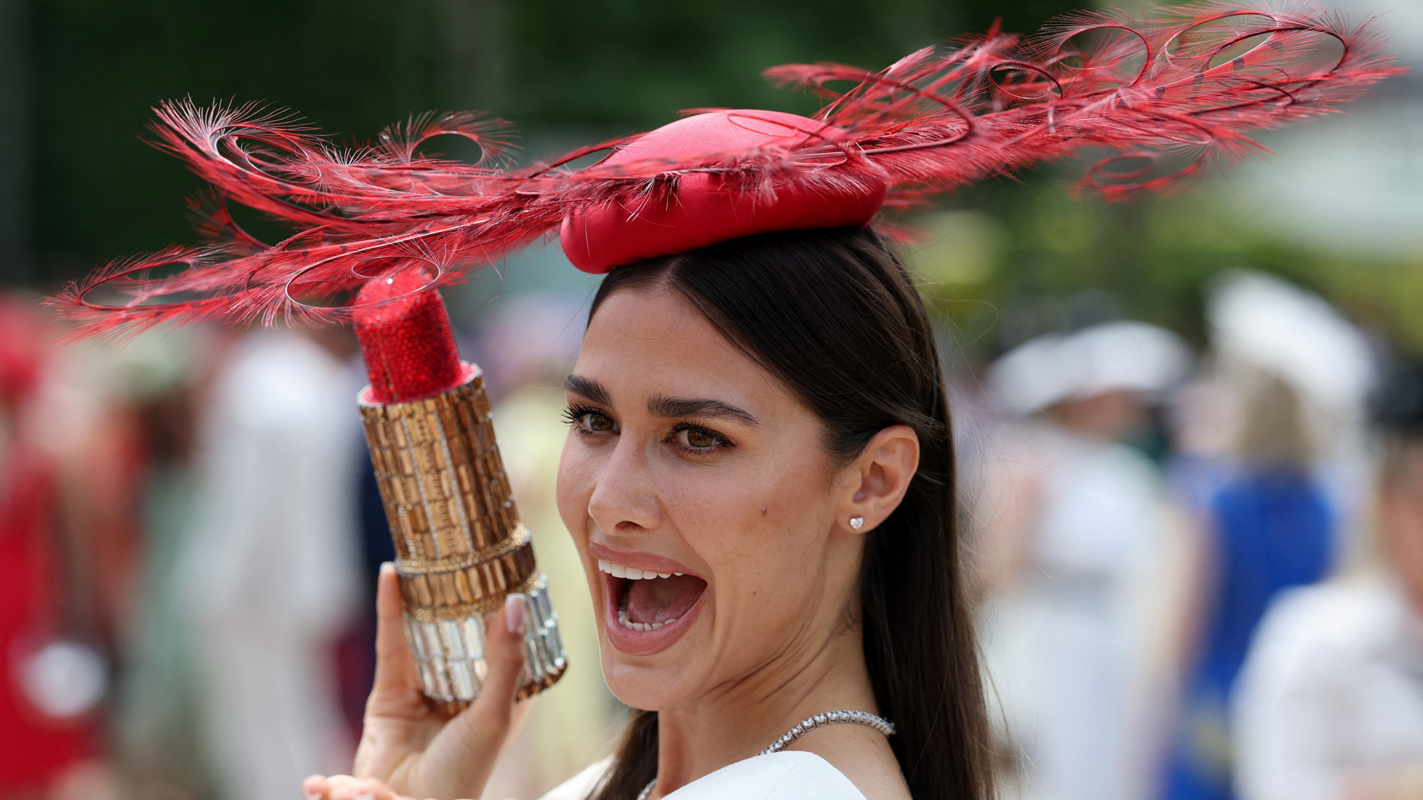 A woman wearing a red fascinator, holding a model of a large lipstick.