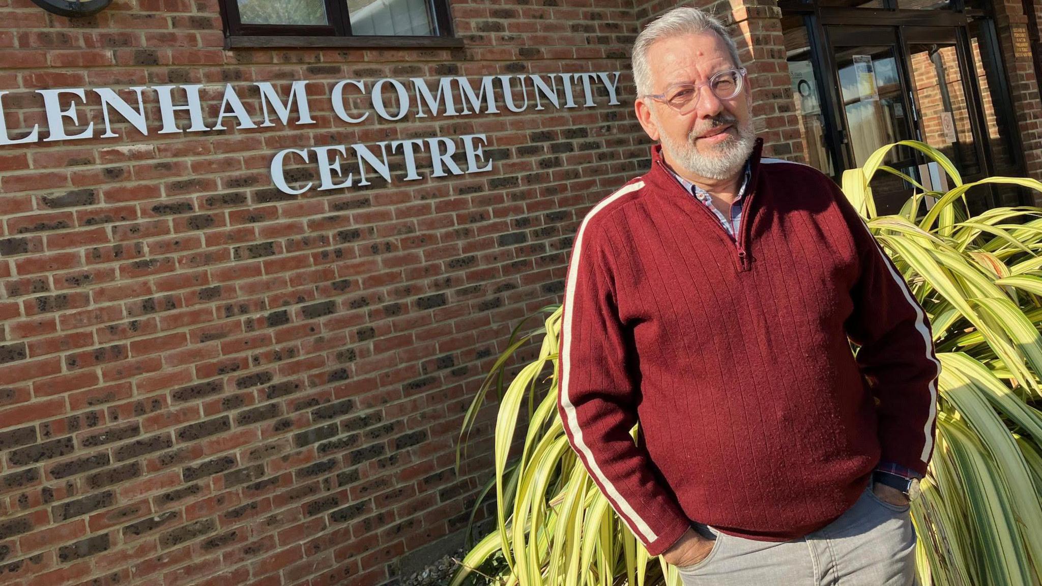 Councillor John Britt stands in front of Lenham Community Centre, wearing a burgundy jumper and beige trousers. He has grey hair and a beard and wears glasses and is standing with his hands in his pockets.