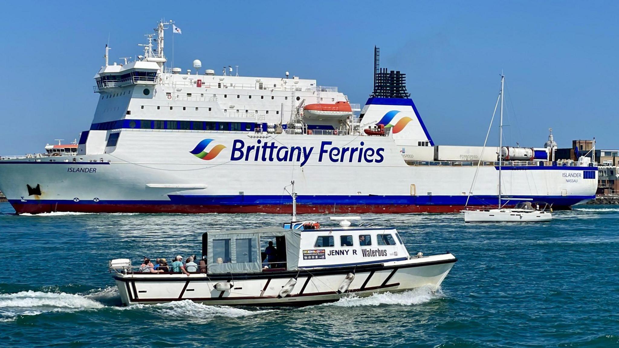 A large ferry is berthed. It is a white bird with the words Brittany Ferries in navy blue along the side. orange lifeboats are visible on the ferry. There is a smaller white boat passing in front of it. A few passengers can be seen on the deck of the smaller vessel.