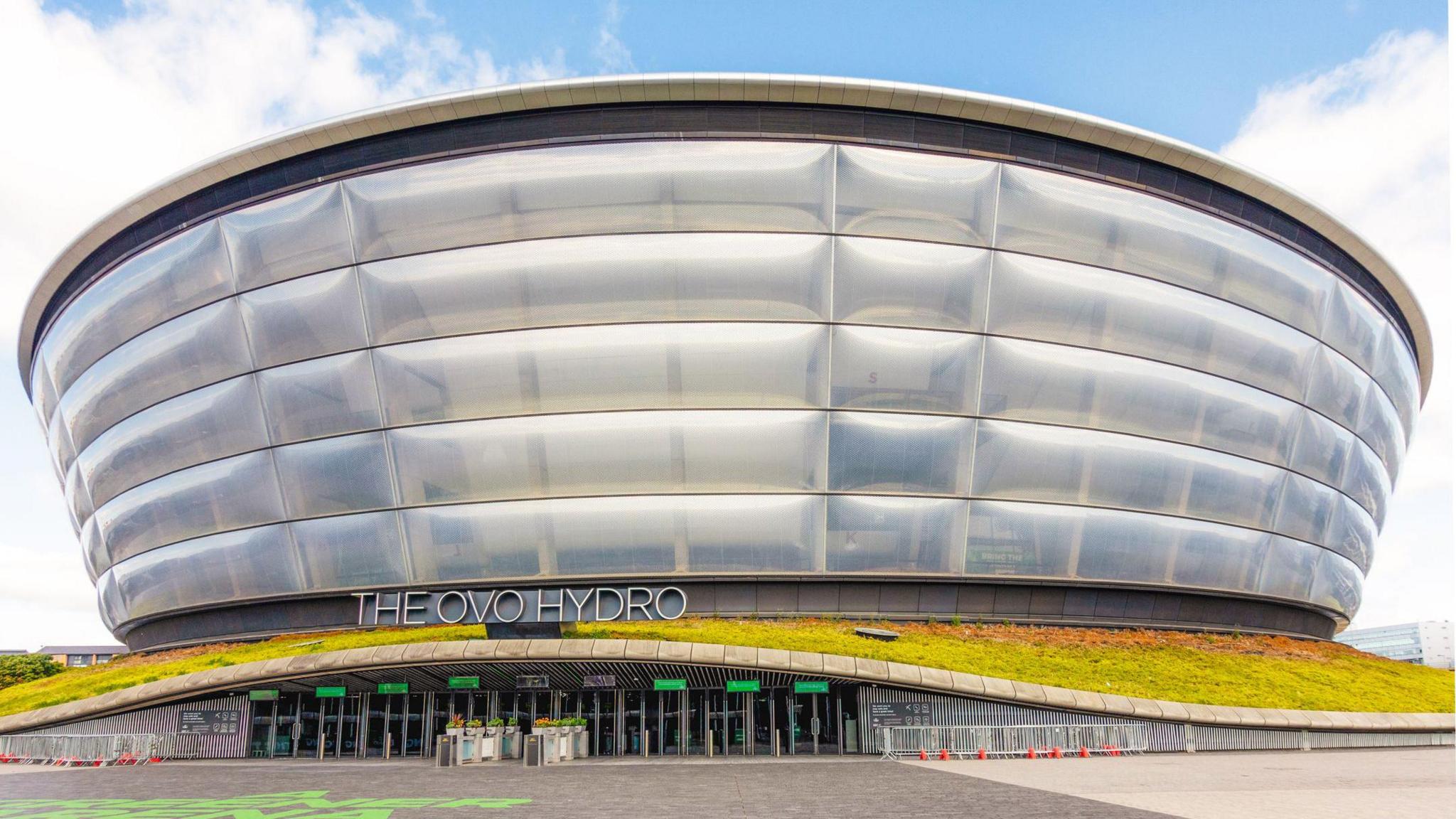 The Ovo Hydro is a tall, round silver building which looks like several large silver bands piled on top of one another. The entrance has several doorways and a sign above the entrance reads THE OVO HYDRO. The entrance has a small band of grass on top of it which snakes around the building. The land approaching the entrance is smooth grey pavement.