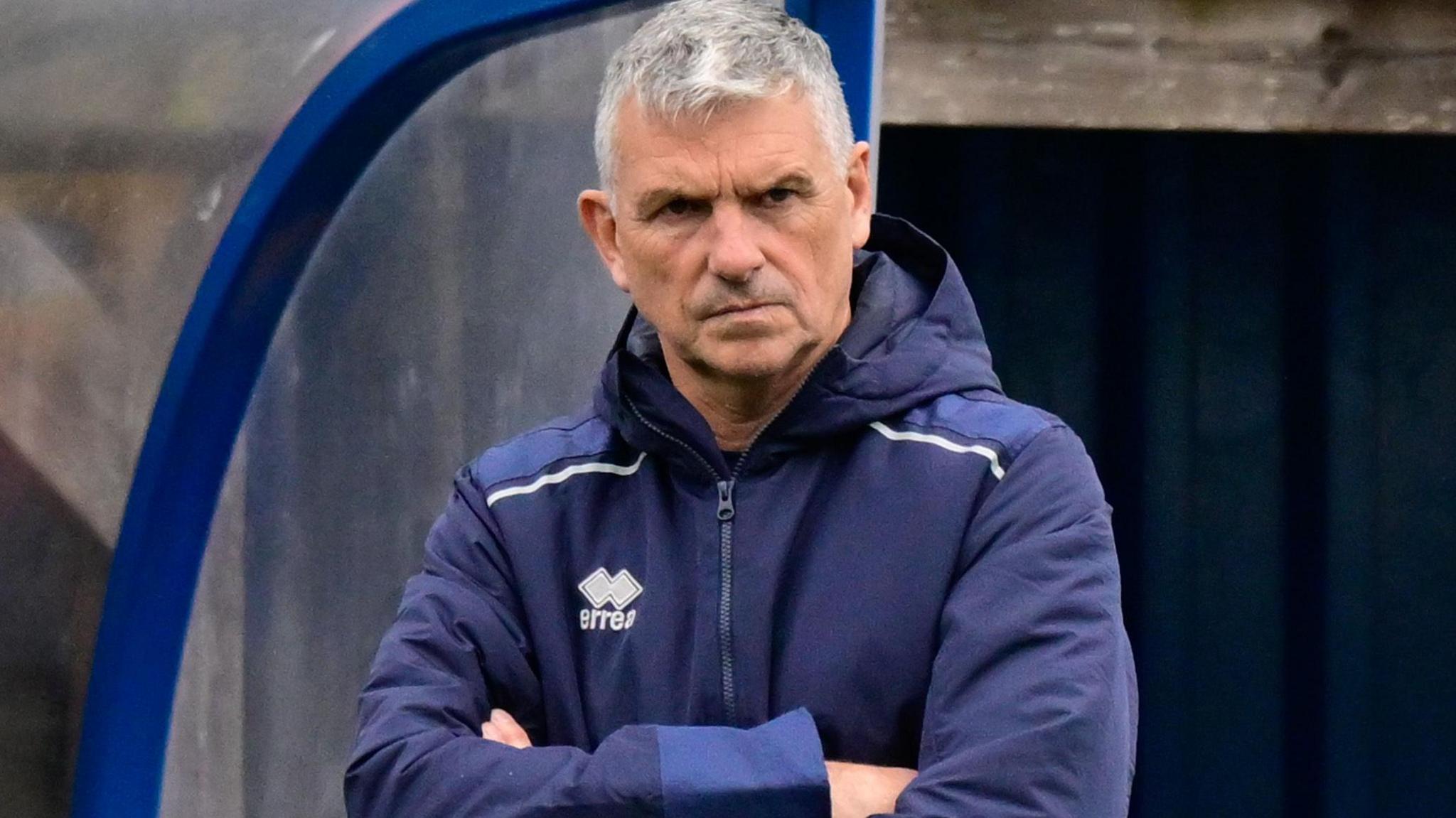 John Askey with his arms crossed while stood in the dugout