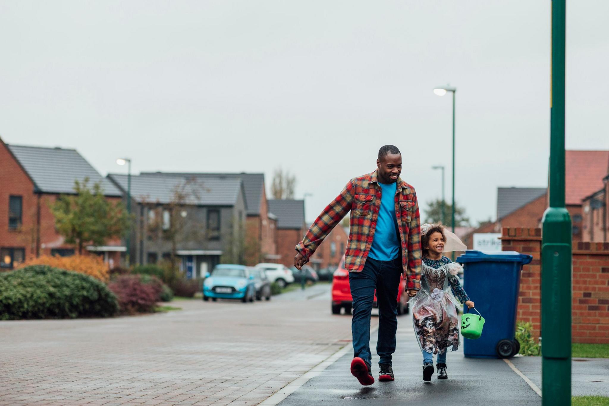 a girl and her dad walking down a UK street. the girl is wearing a spooky costume and holding a bucket for sweets. they are smiling and holding hands.