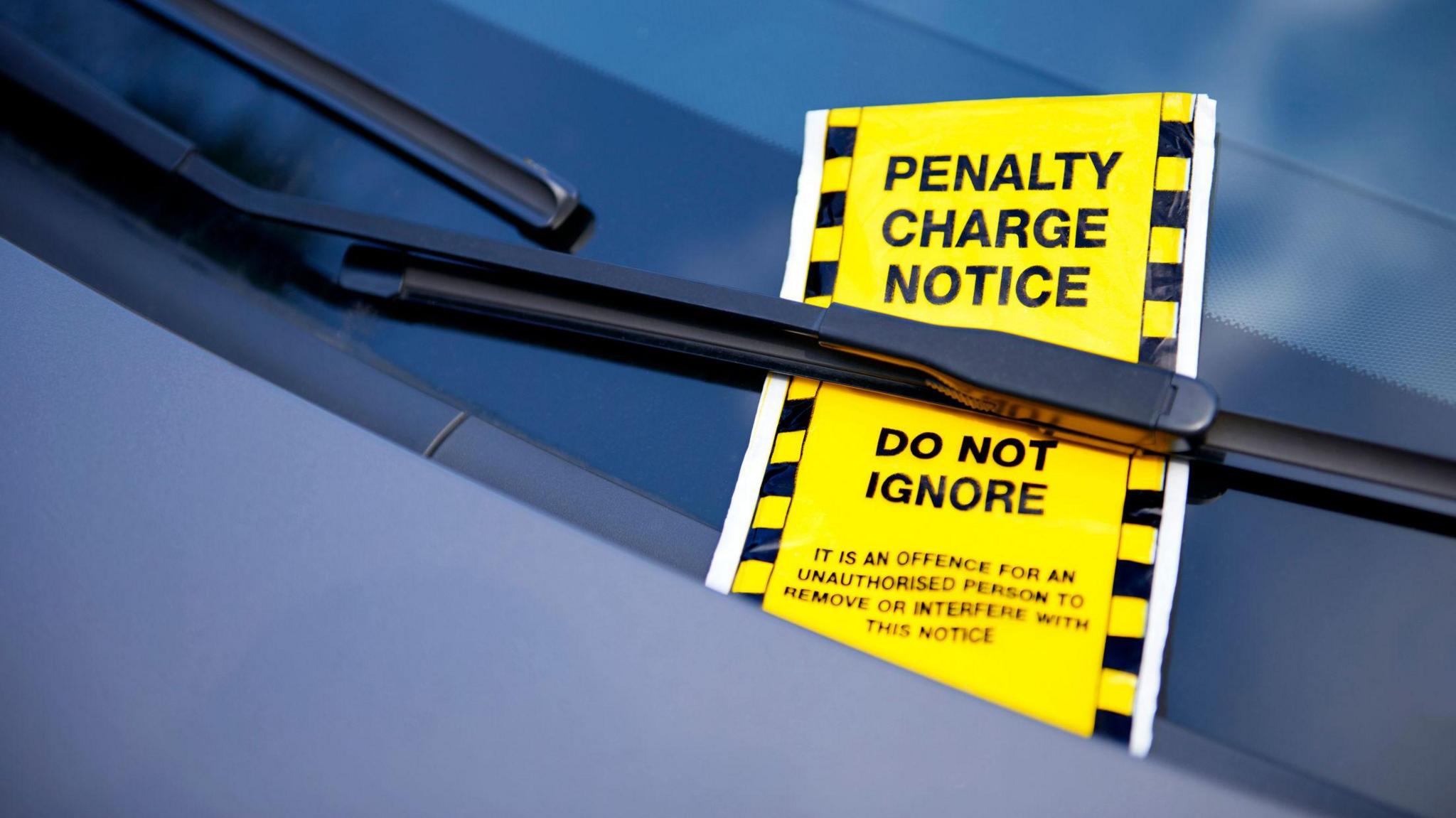 A close-up image of a yellow penalty charge notice placed under the windshield wiper of a car. The notice has bold black text reading "PENALTY CHARGE NOTICE" and "DO NOT IGNORE," with a warning about unauthorised removal.