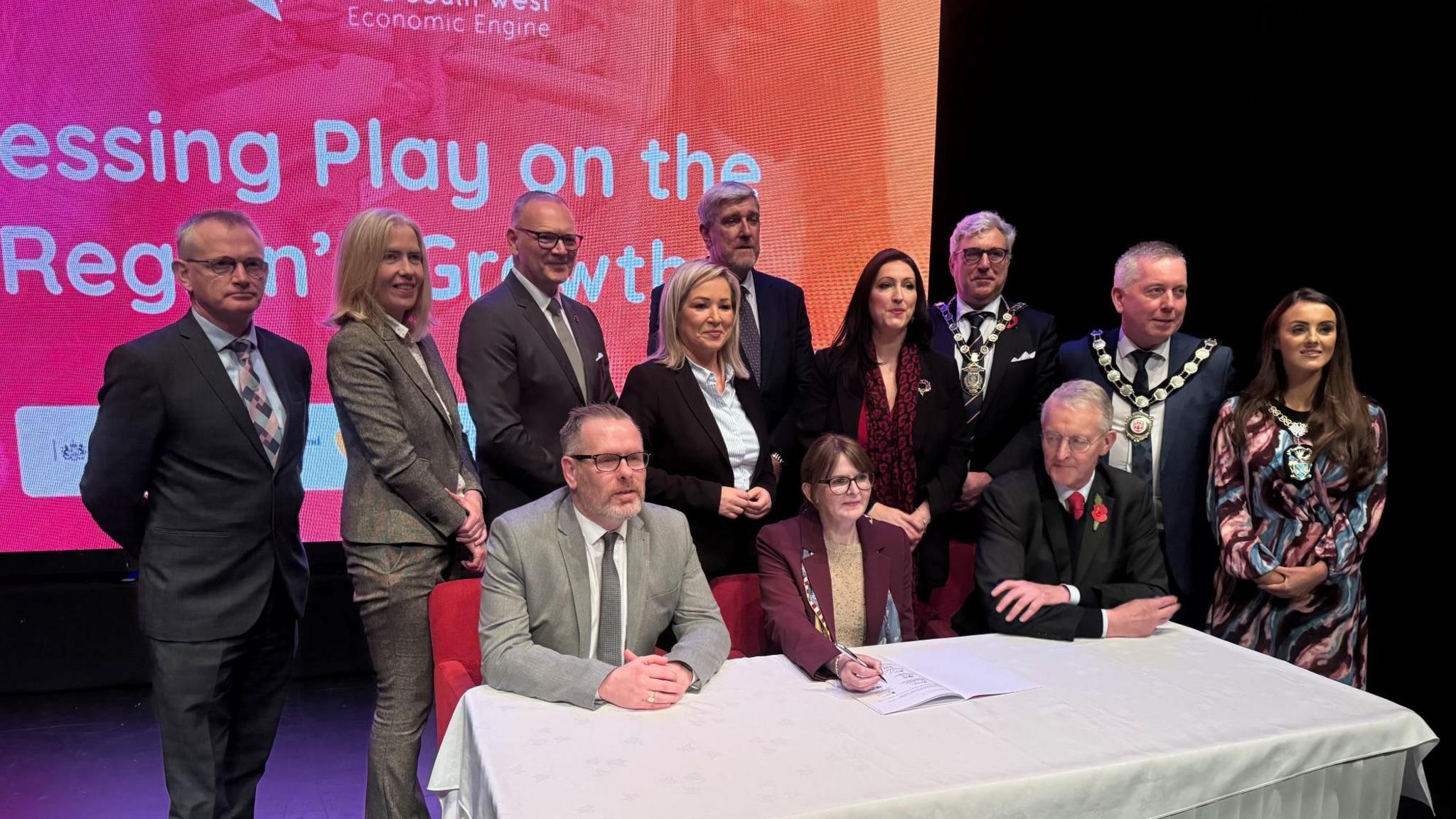 12 men and women gather round a table, with a white cloth covering it. 
Three are seated at the table, a women wearing a burgundy blazer and sand coloured t-shirt, sits inbetween two men with pen in hand ready to sign the paper. 