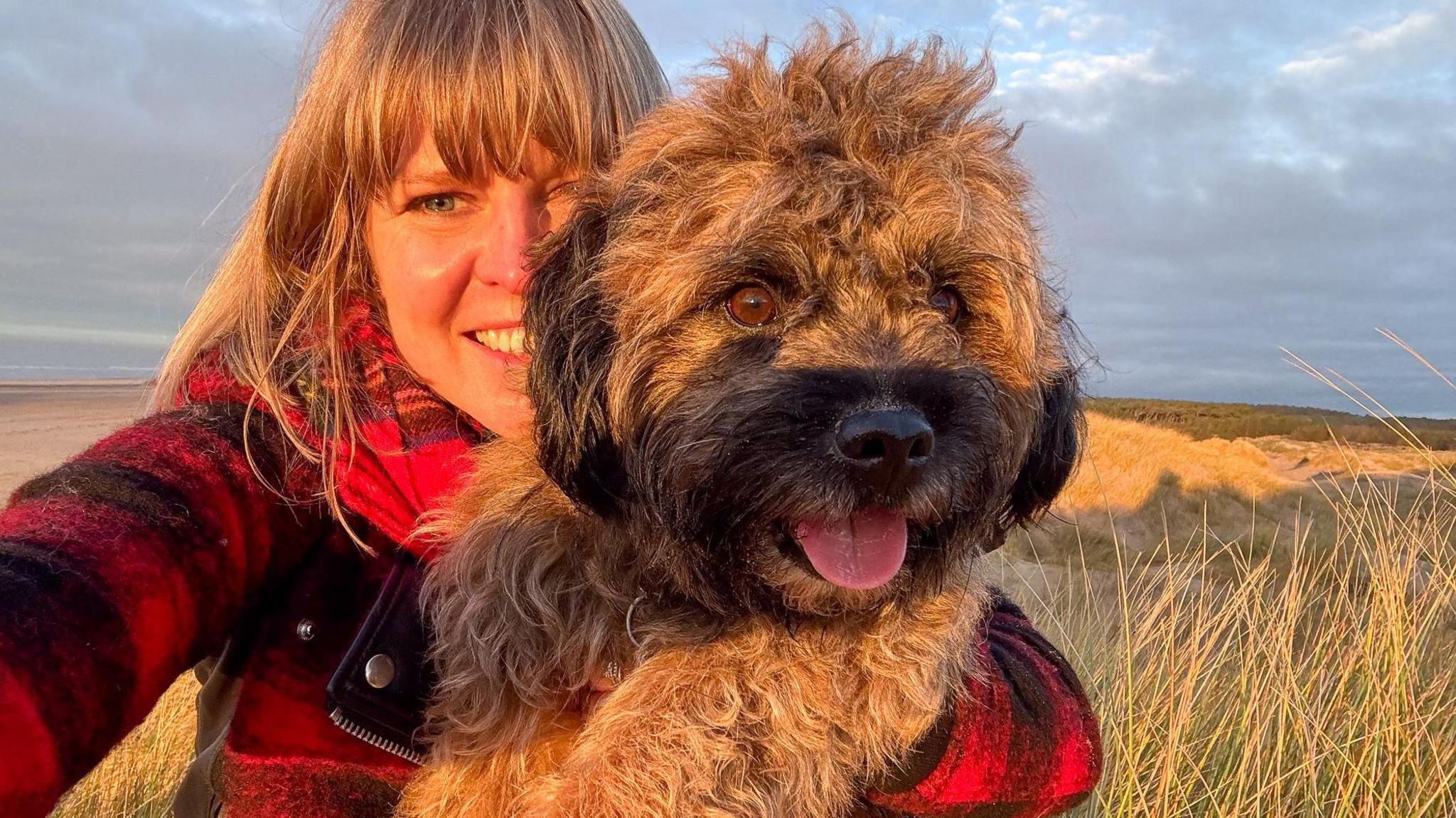 A woman in a red jacket cuddles a small, dog. The dog is looking into the distance and has its tongue out and looks happy. There is a beach in the background and it is a sunny day. 