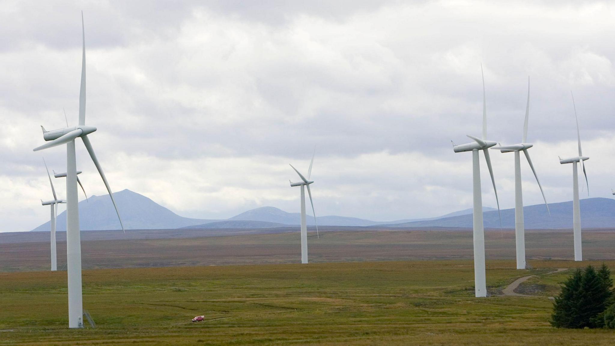 Seven grey turbines on a wide open space of peat and moorland. In the distance are hills and mountains.