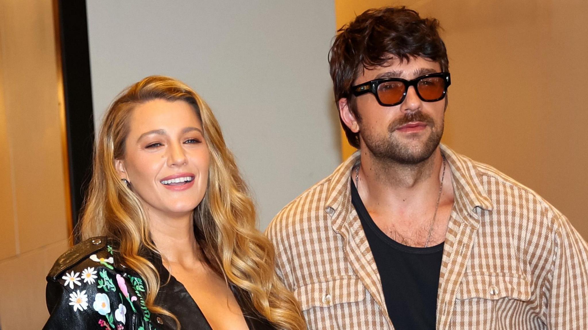 Blake Lively and Brandon Sklenar pictured in New York in August. Blake is a woman in her 30s with long curled blonde hair. She wears a painted leather jacket and smiles. Brandon, a 34-year-old man, has shaggy brown hair and a short stubbled beard. She wears brown tinted sunglasses and a brown gingham shirt over a black vest. 