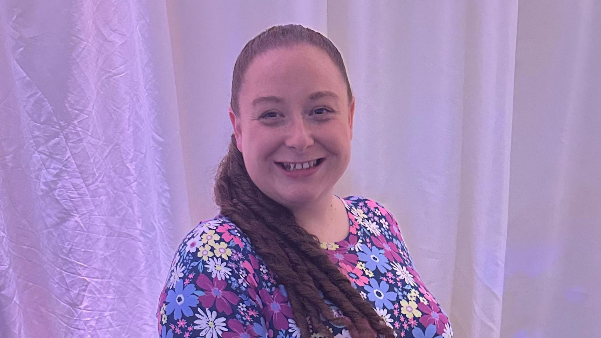 Leah Clapham smiles at the camera. She has long brown hair that is tied up and hangs over her shoulder. She wears a floral dress.
