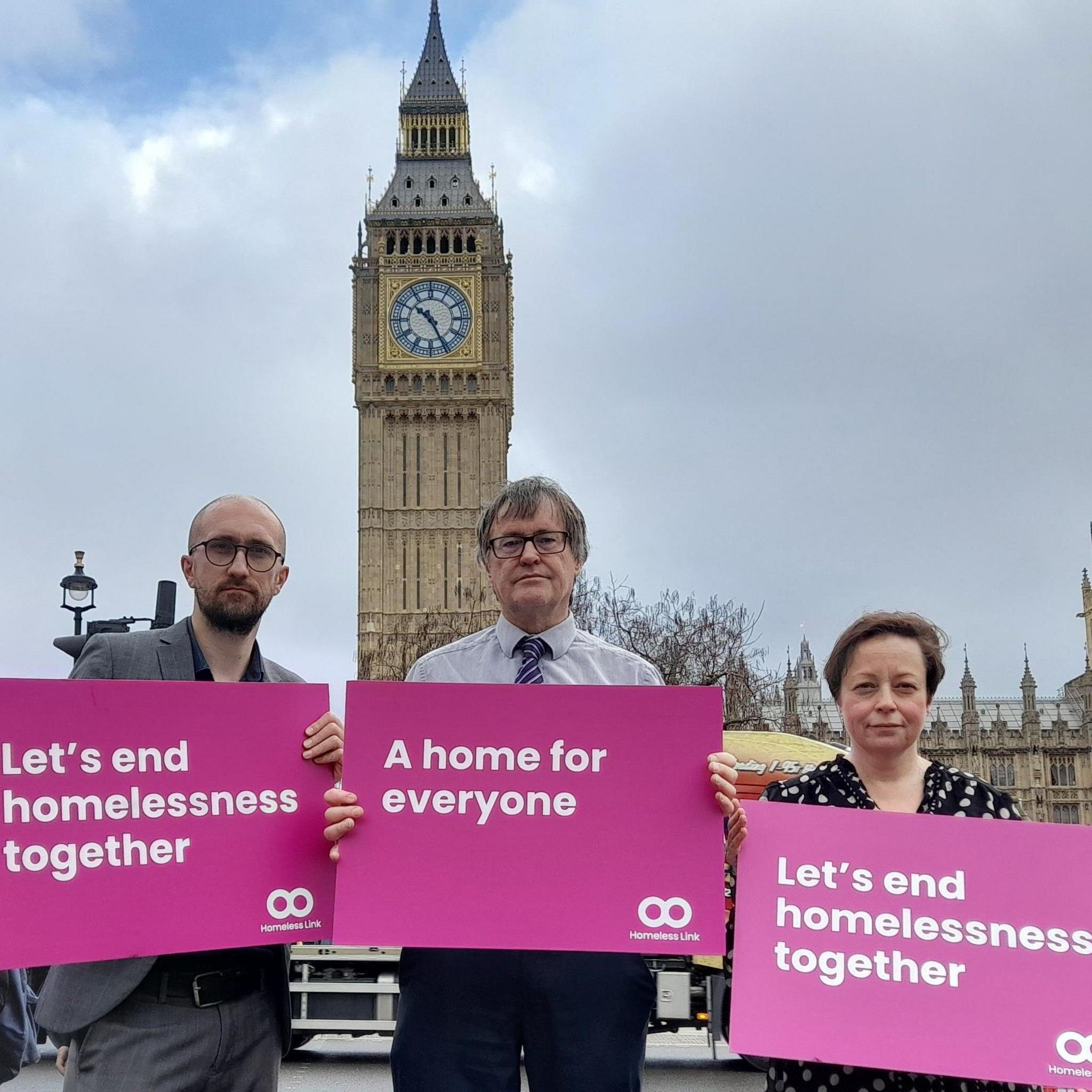 Three people are standing in front of Big Ben holding placards that say "Let's end homelessness together" and "A home for everyone"