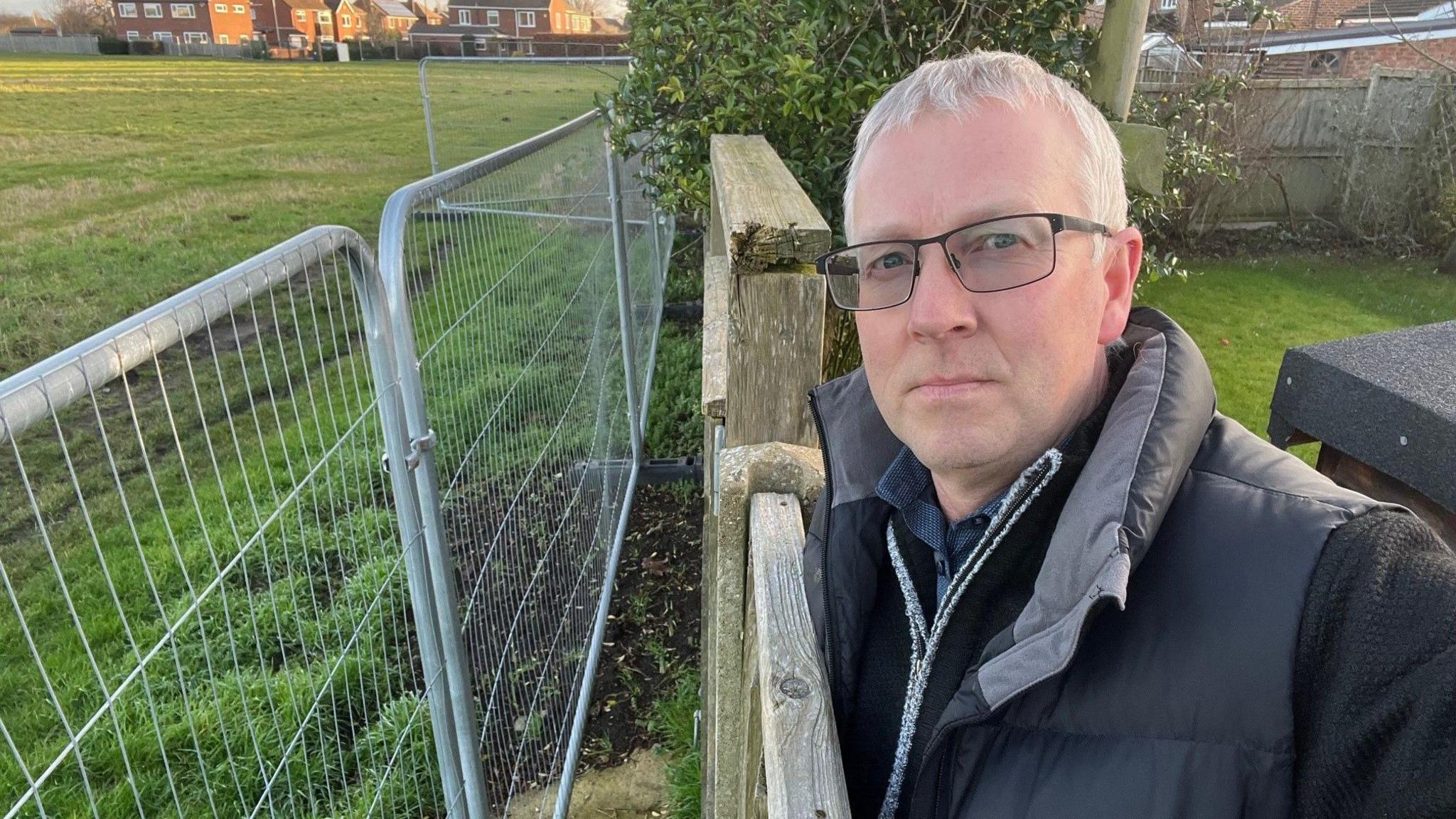 Mark Palmer wearing a black coat and black framed glasses. He is standing at the end of his back garden over looking the field, where metal fencing has been put up just the others side of his wooden fence.