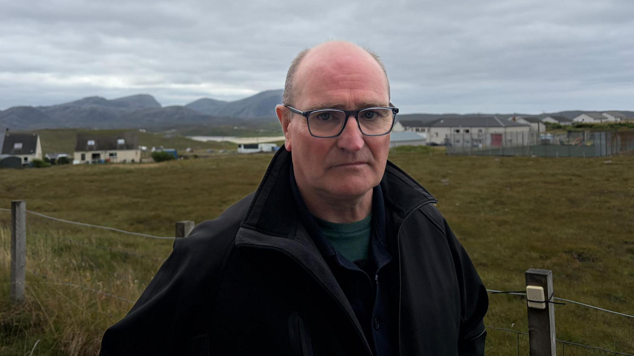 Fear maol le seacaid dhubh air agus speuclairean. Tha e na sheasamh a-muigh le togalaichean geala air a chùlaibh agus beantan fada às. 