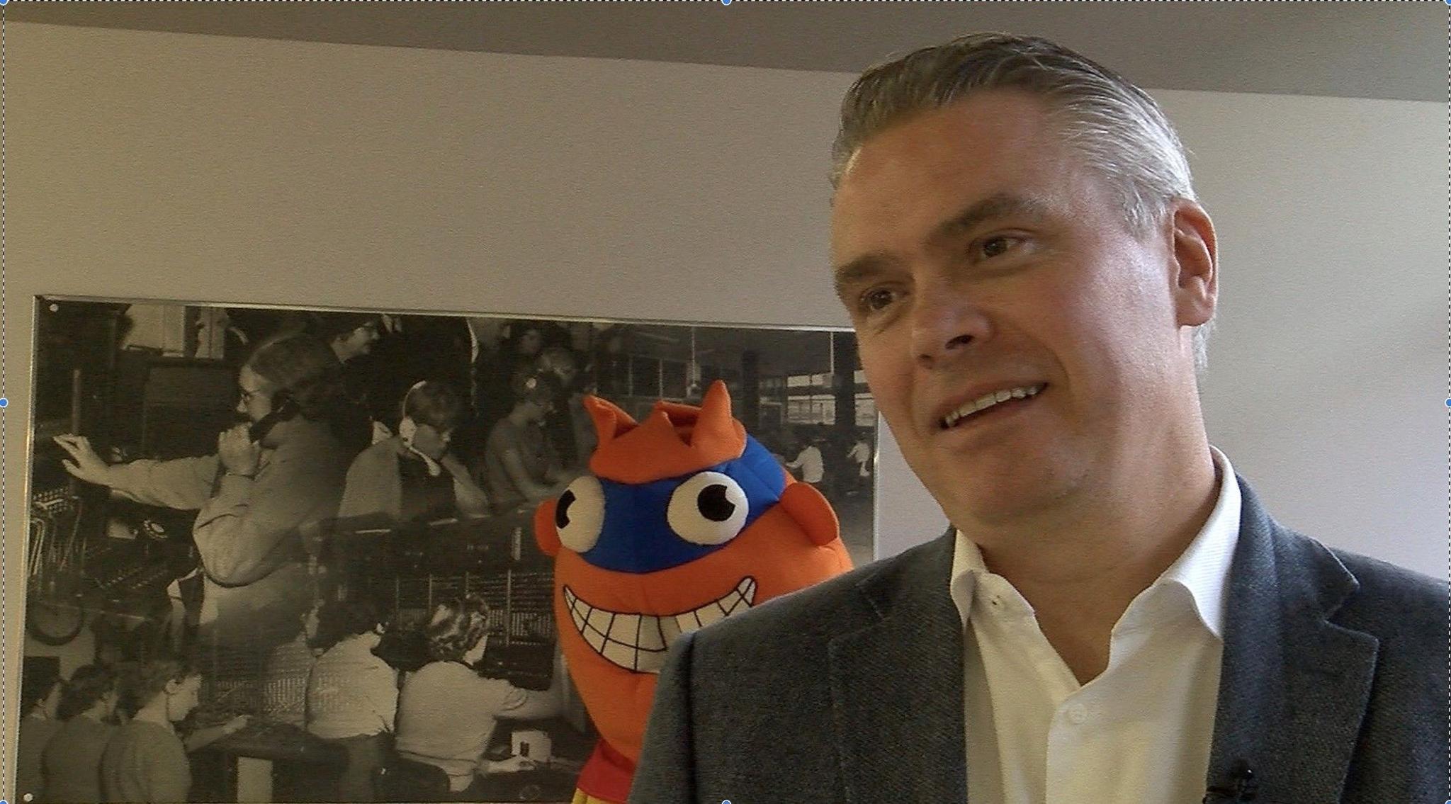 Tim Shaw stands in front of a large blue and orange plush toy and a black-and-white canvas showing scenes from KCOM's history. He has short grey hair, slicked back, and wears a smart white shirt and grey jacket.