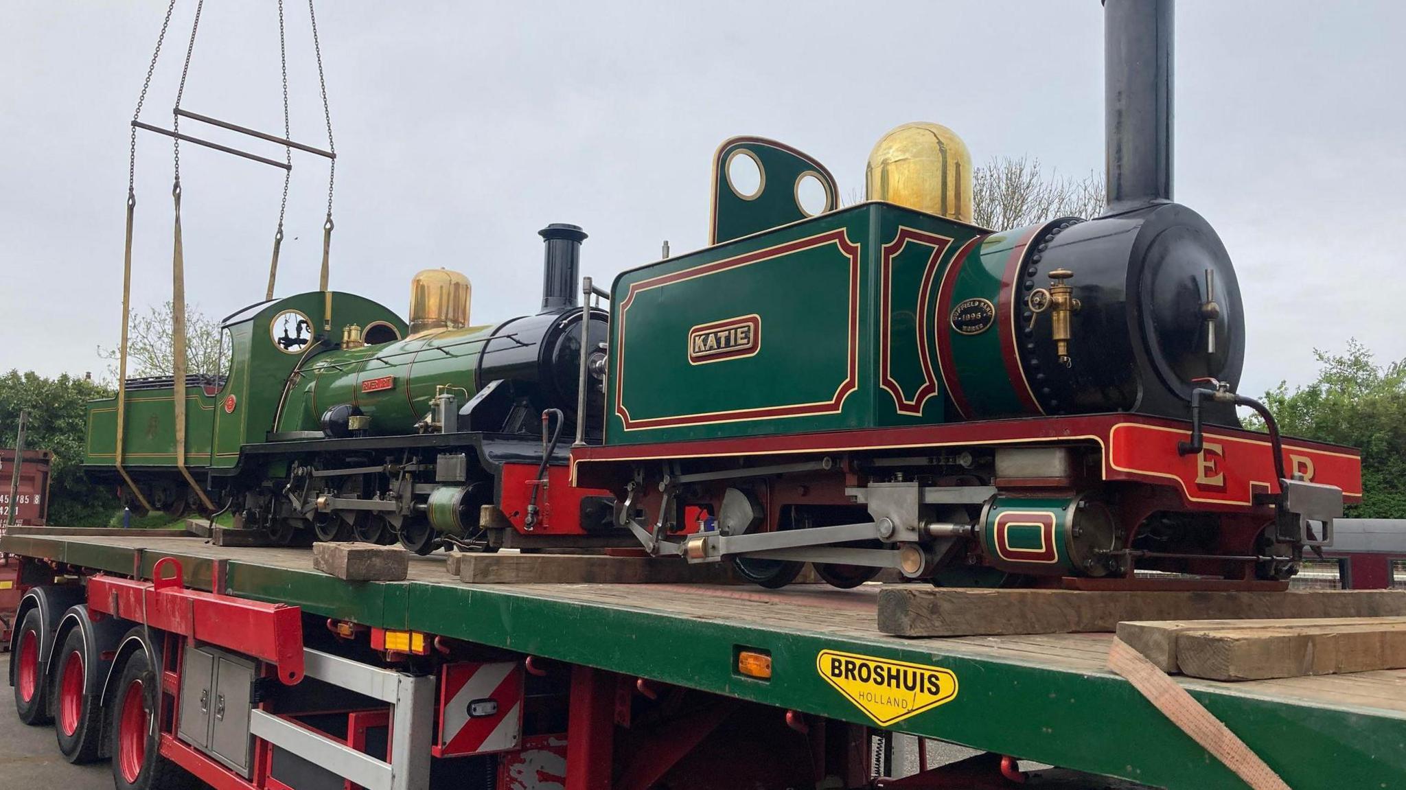 Two small-size steam locomotives on a large trailer. They are dark green with black funnels, and the one in the front is named Katie