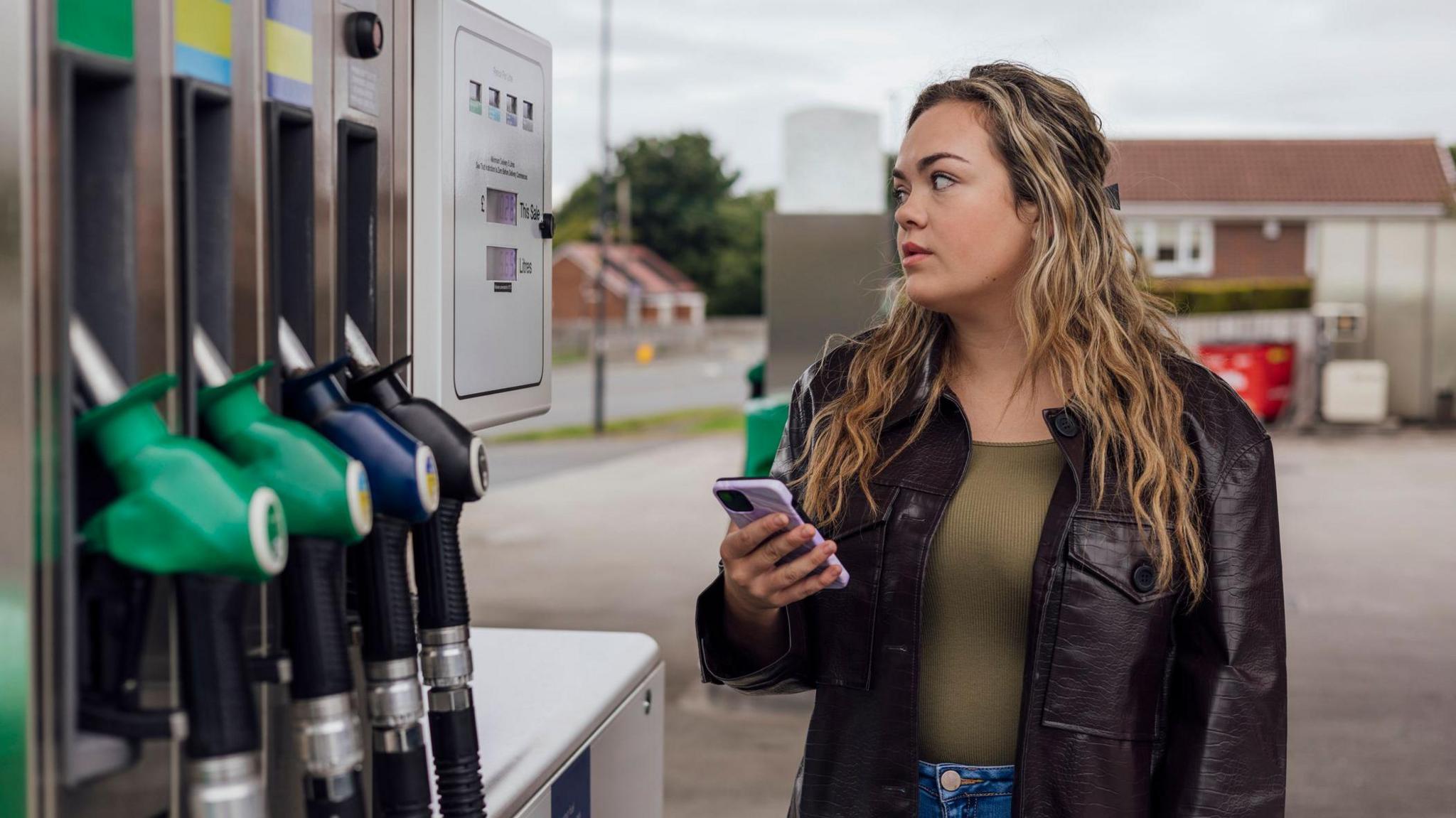 Woman holding a phone filling up a car at a petrol station - stock image