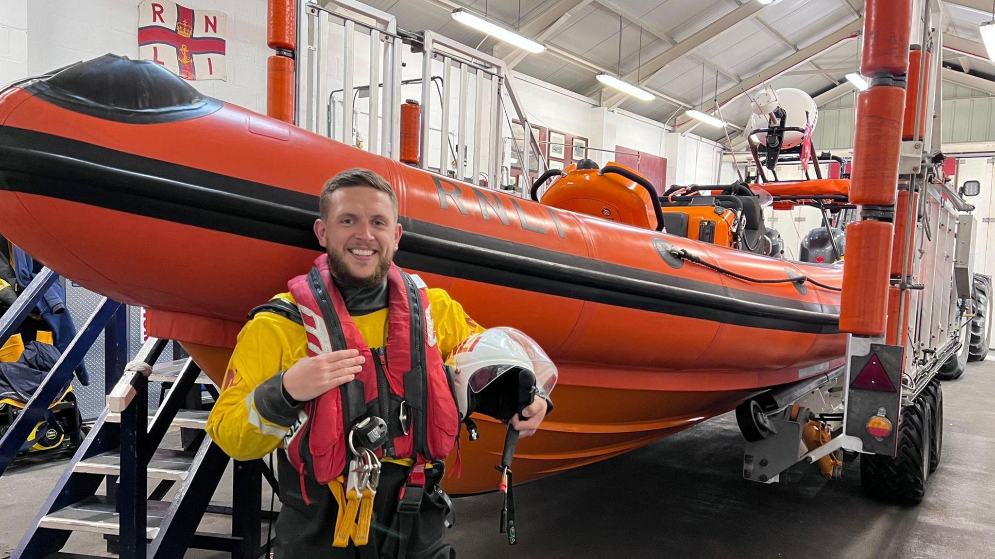 Connor Wray in his yellow lifeboat volunteer uniform and red lifebelt, holding a helmt and standing in front of an elevated orange RNLI boat in its station depot