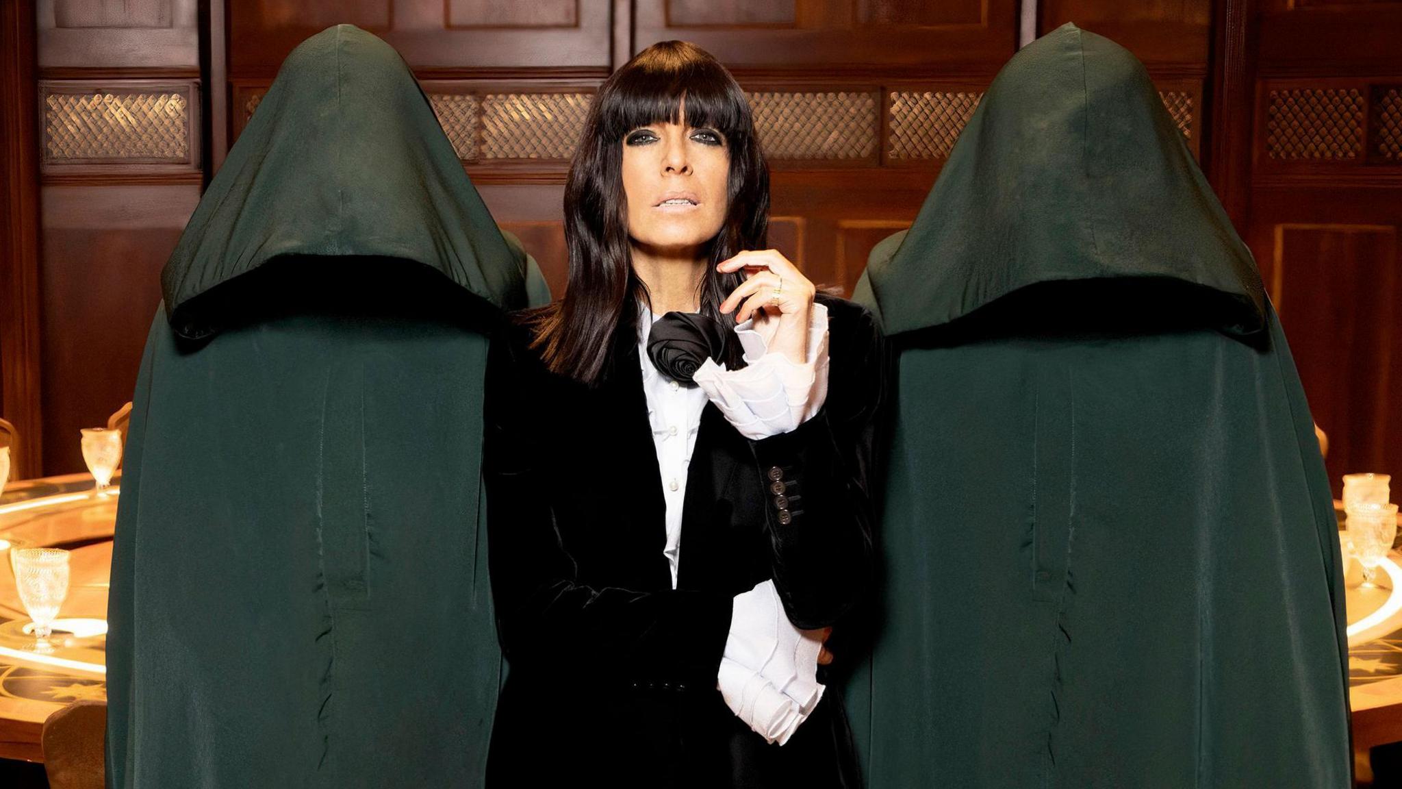 Claudia Winkleman stands in front of the round table on the set of The Traitors - wearing a black jacket with white shirt and ruffled sleeves - with long dark hair, on either side there are two people wearing black cloaks and hoods hiding their faces.