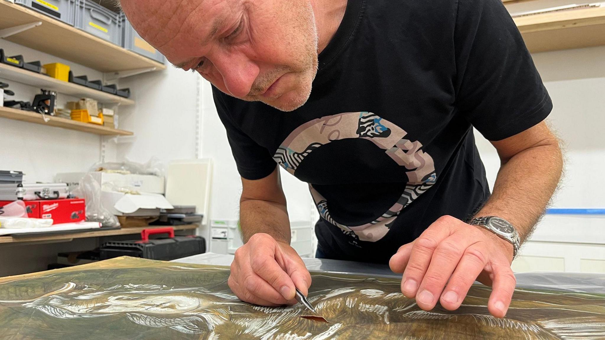 A man in a black T shirt leans over a large artwork fixing a slit in the paper