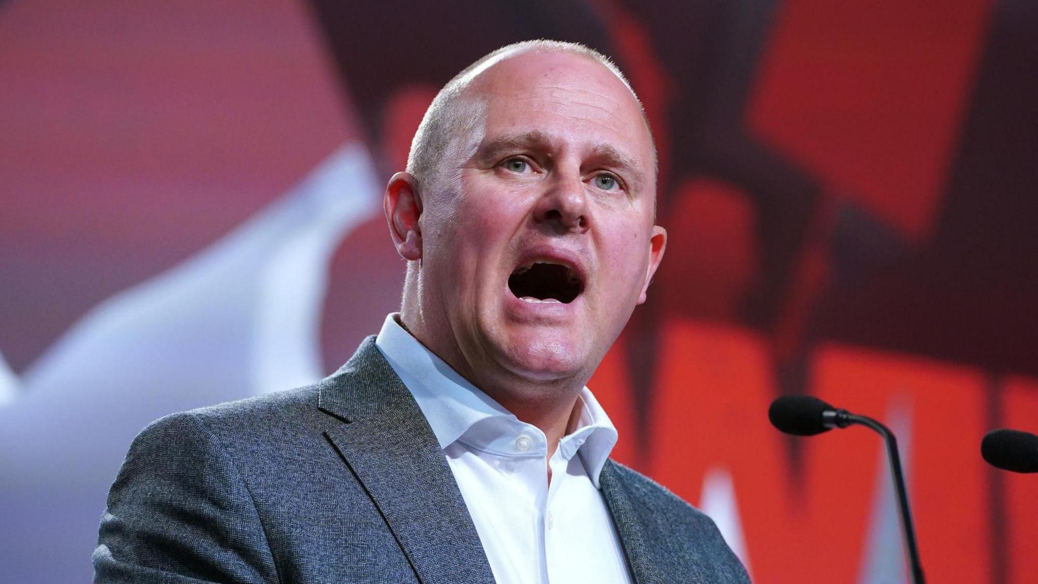 Paul Nowak, wearing a grey suit and white shirt, speaks from a podium with a red wall behind him.